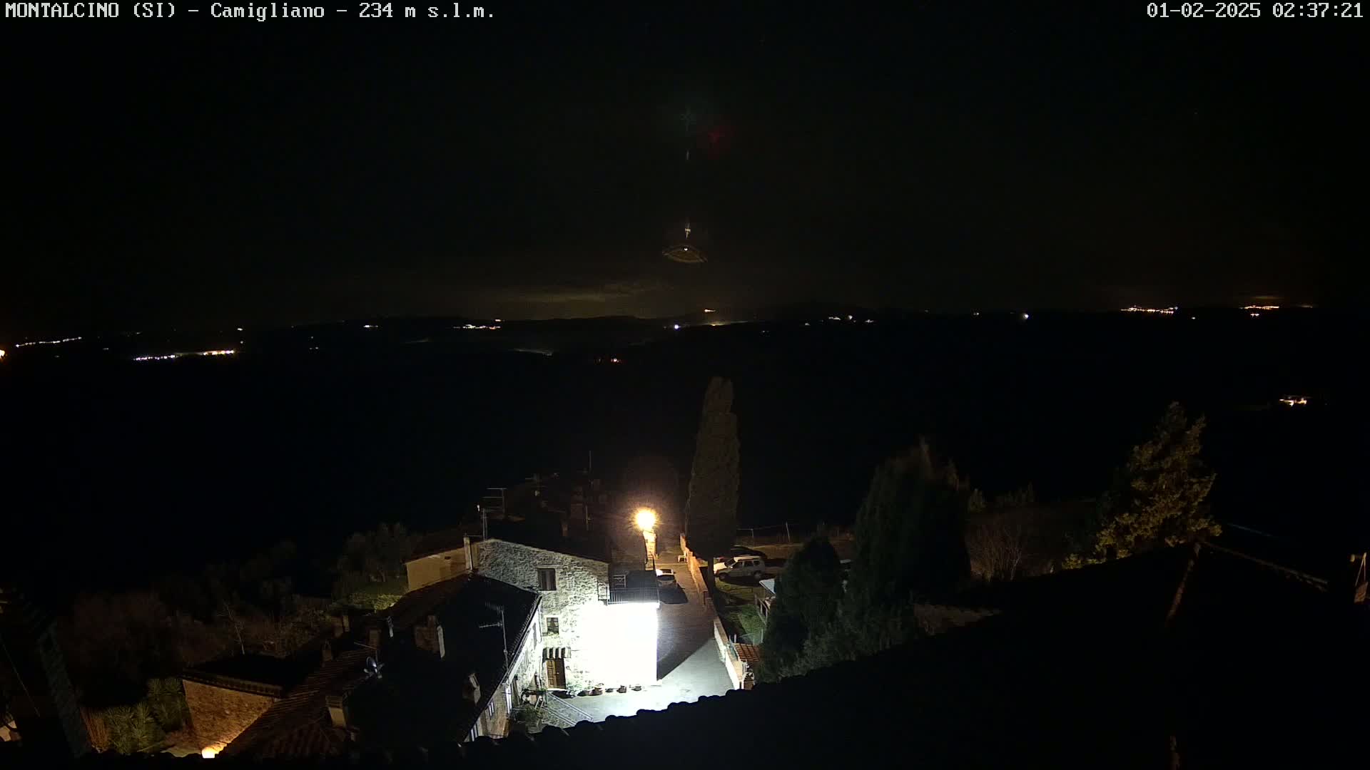 A nighttime view of a small village illuminated by a single bright light, overlooking a dark landscape dotted with distant lights under a mostly clear night sky with a faint, reddish light in the distance.