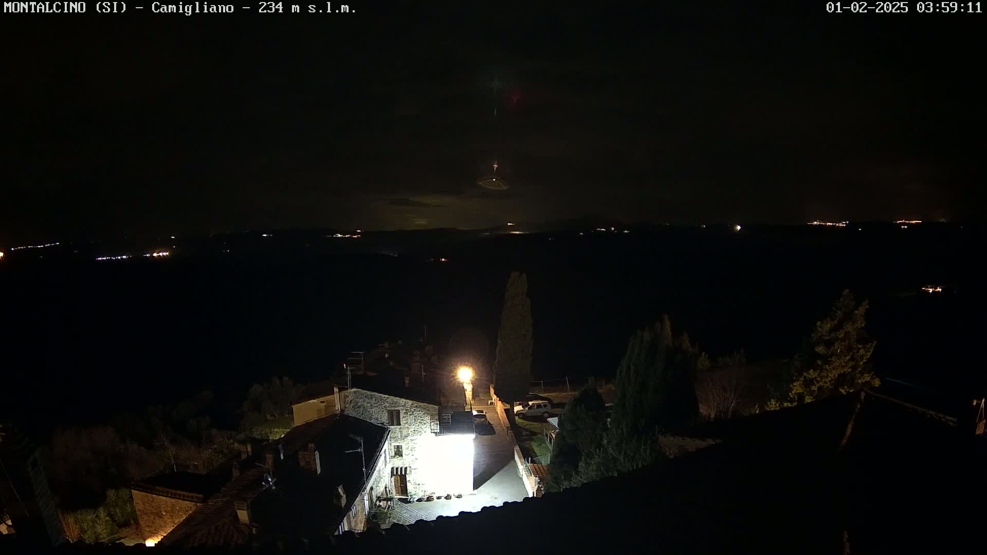 A nighttime, aerial view of a small village with a brightly lit courtyard, nestled in a dark landscape under a mostly clear sky with faint lights visible in the distance.