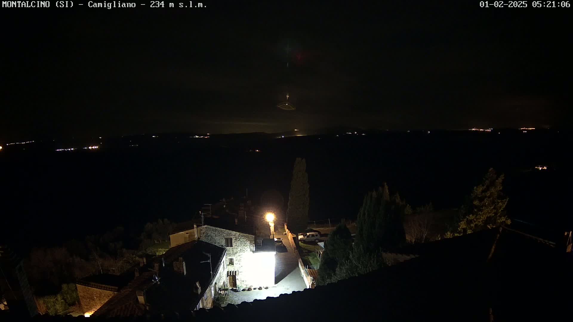 A nighttime view of a village, illuminated by a single bright light, overlooking a dark, distant landscape with a few faint lights visible under a clear, dark sky.