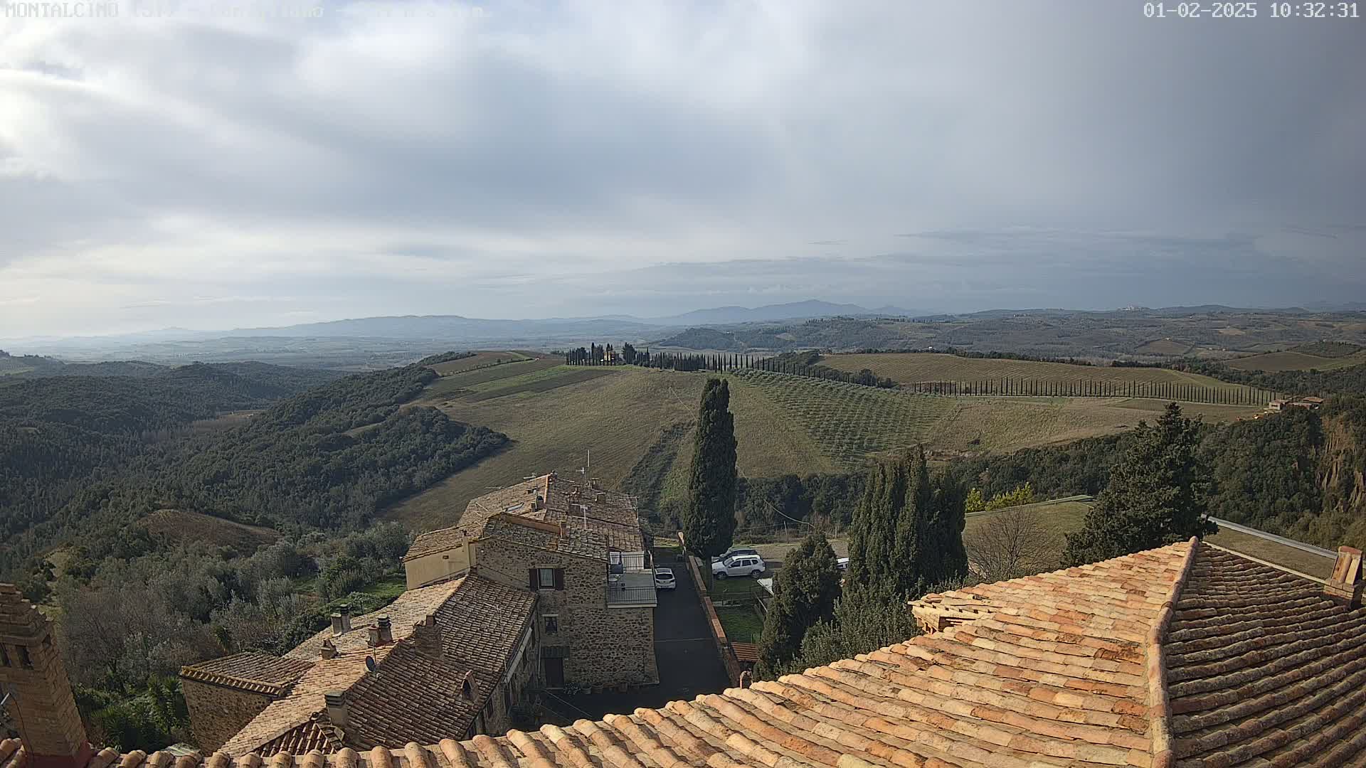 A partly cloudy sky overlooks a hilly landscape of olive groves and stone buildings with terracotta roofs.
