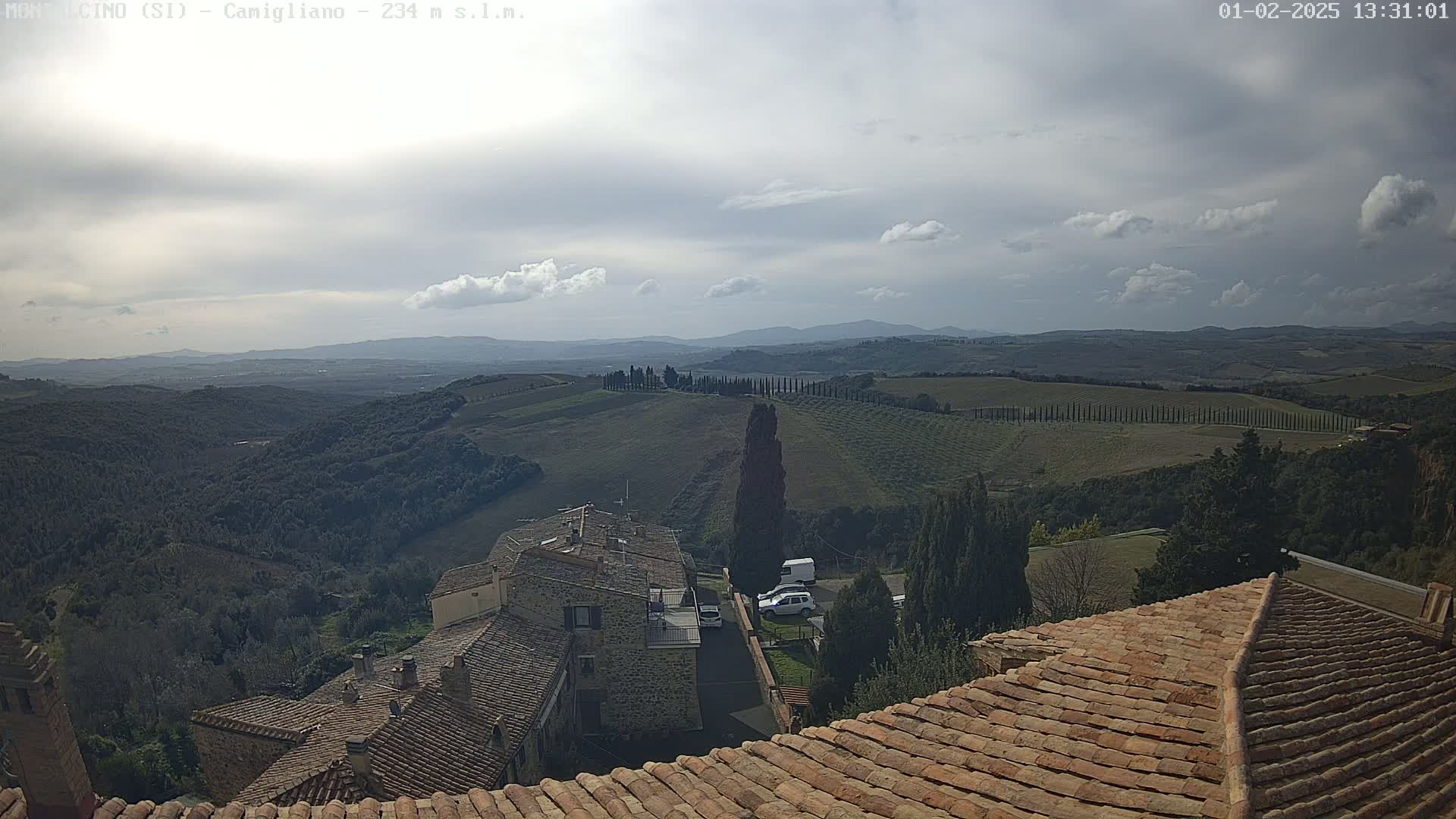 A view from a tiled roof shows a partly cloudy sky over rolling hills, farmland, and a small village.