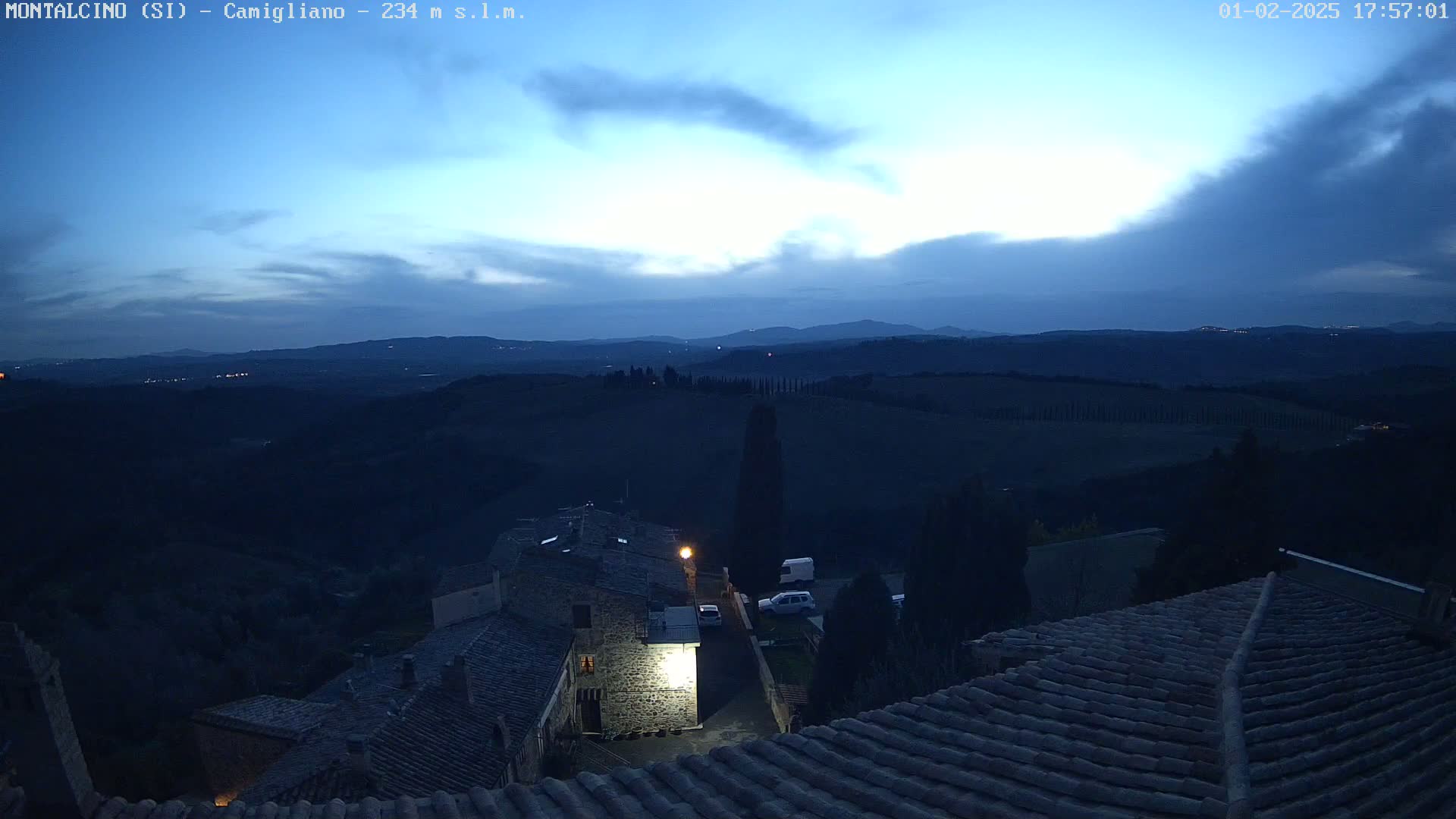 A twilight view from a rooftop shows a small village nestled in rolling hills under a mostly clear, dark blue sky.