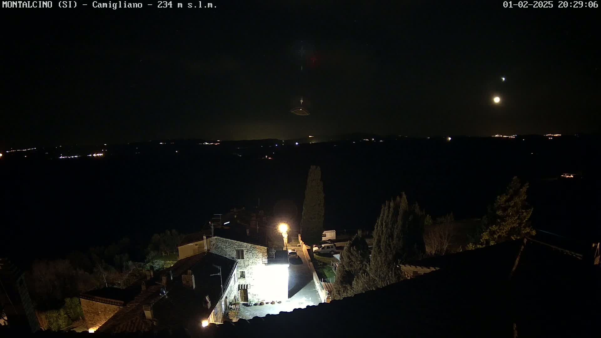 A nighttime, high-angle view shows a small village illuminated by streetlights, under a dark sky with a bright moon and what appears to be a meteor or other atmospheric phenomenon.