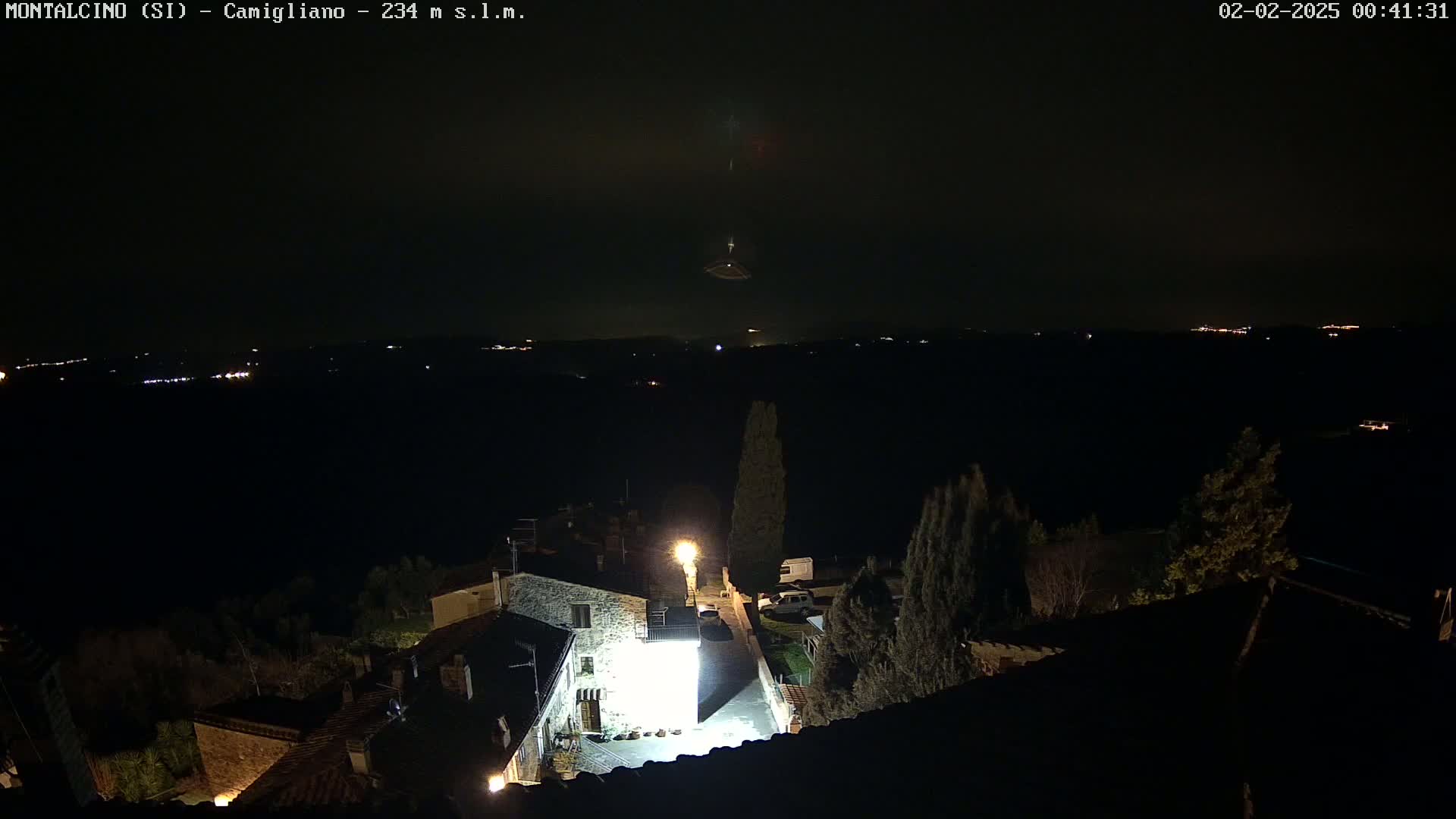 A nighttime view of a village illuminated by a few lights, with a distant, hazy horizon featuring faint lights and a dark, possibly cloudy sky.
