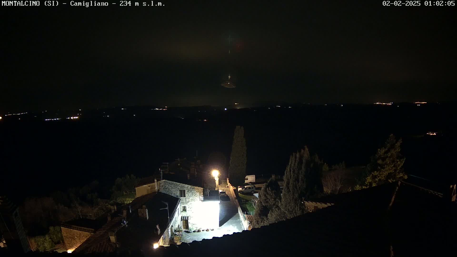 A nighttime view of a small village illuminated by artificial light, overlooking a dark, distant landscape under a mostly clear sky.