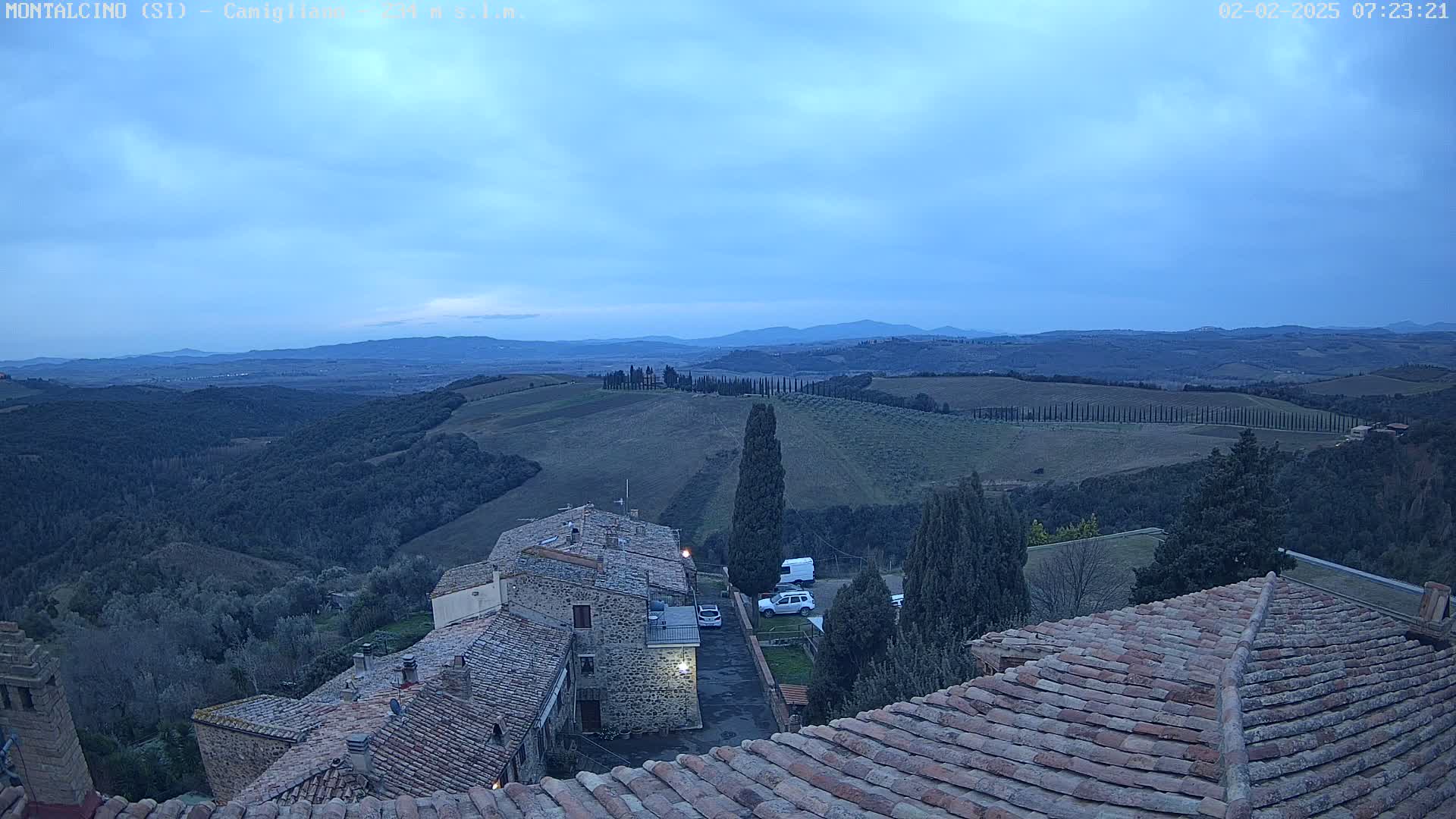A twilight view shows rolling hills dotted with cypress trees and stone buildings with terracotta roofs, under a mostly clear, dusky blue sky.