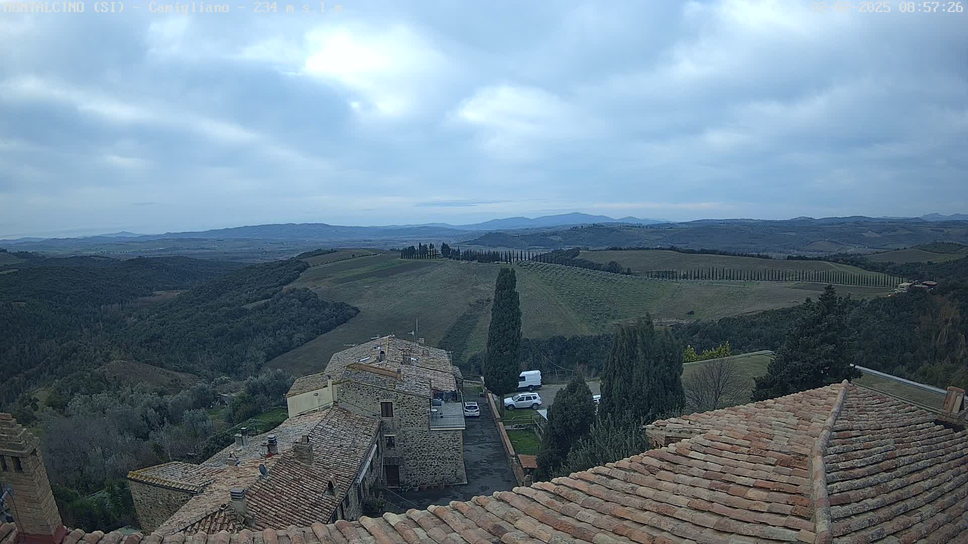 Stone buildings with terracotta roofs overlook a hilly landscape of olive groves and cypress trees under a cloudy sky.