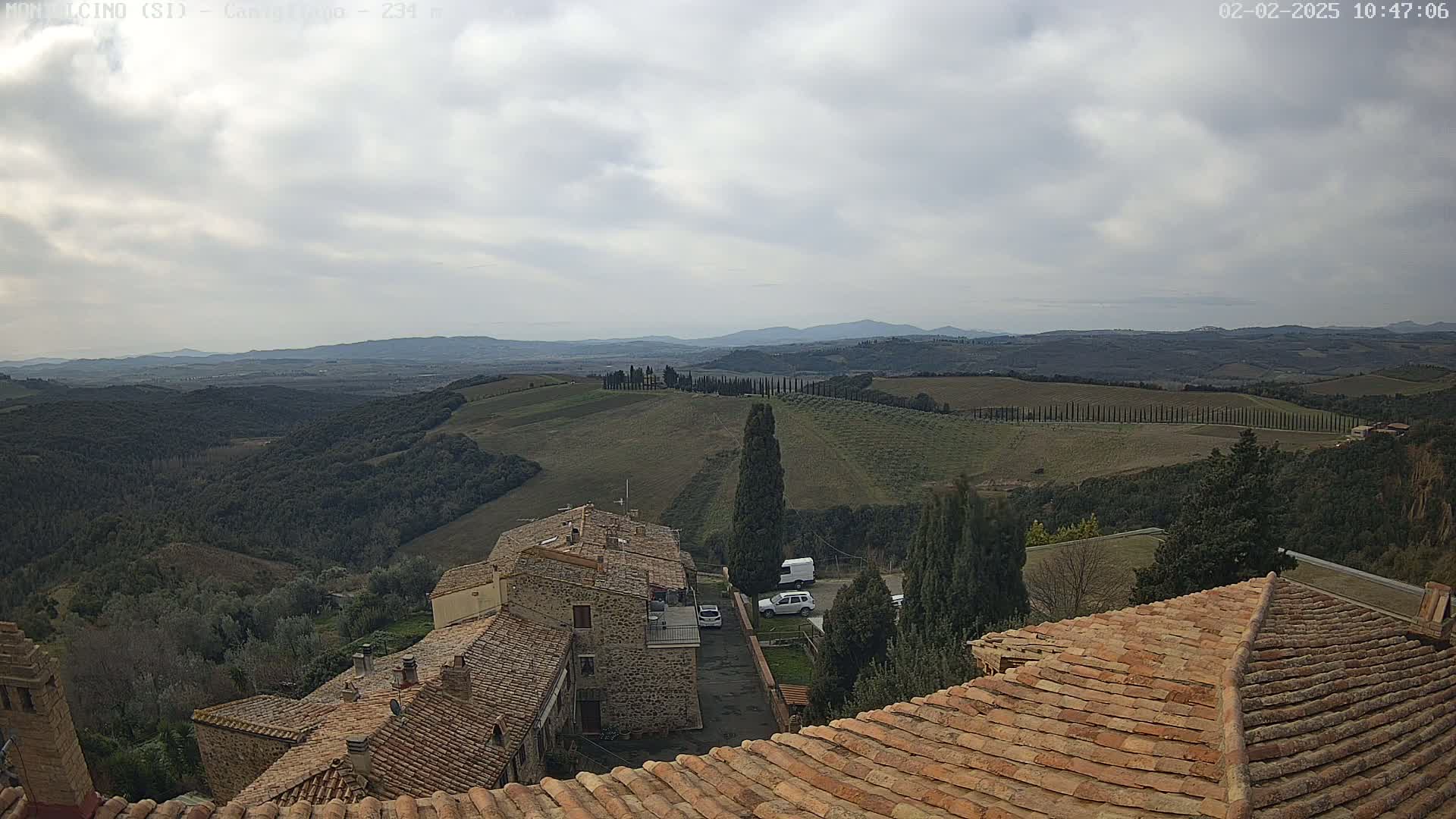 Under a cloudy sky, terracotta rooftops overlook a hilly landscape dotted with cypress trees and stone buildings.