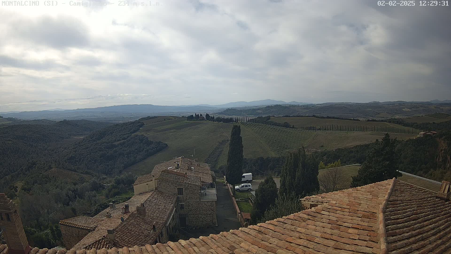 A view from a tiled rooftop shows a cloudy sky over rolling hills dotted with trees and buildings, including a cluster of stone houses.