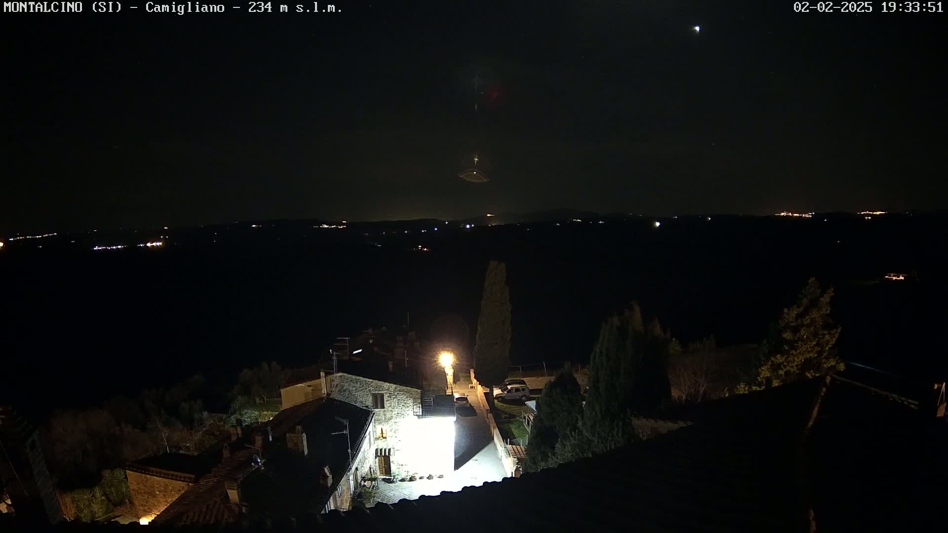 A nighttime view of a village illuminated by streetlights, with a dark, starlit sky and distant lights on a hillside.