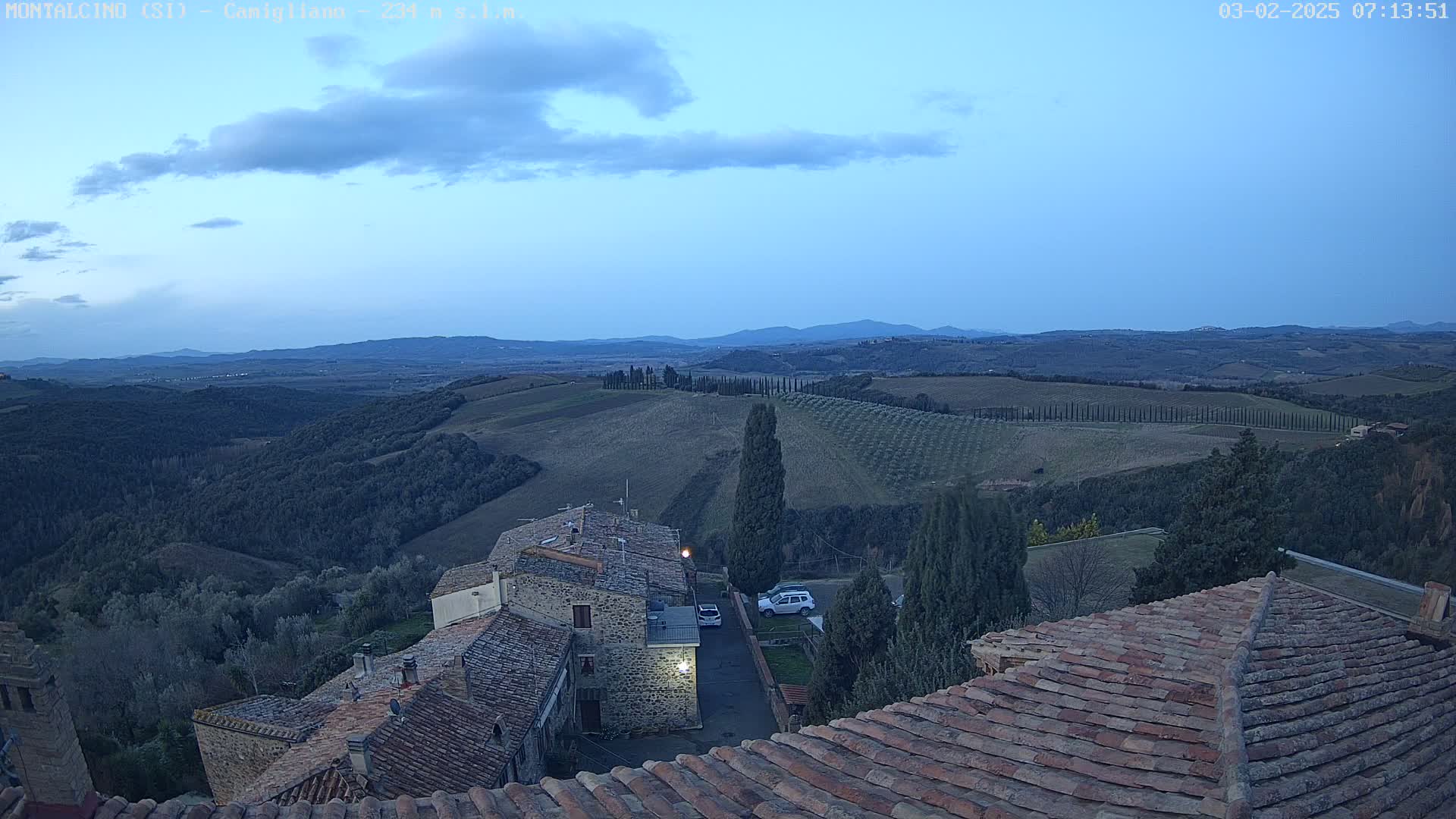 A view from a rooftop shows a twilight landscape of rolling hills, olive groves, and a small village under a partly cloudy sky.