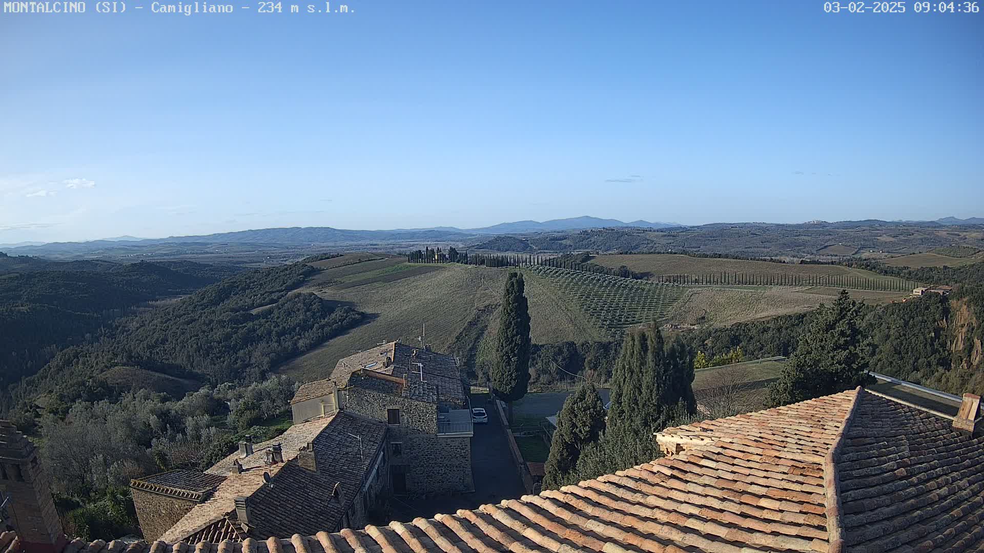 A sunlit view of rolling hills dotted with trees and buildings, overlooking a village with terracotta rooftops under a clear blue sky.