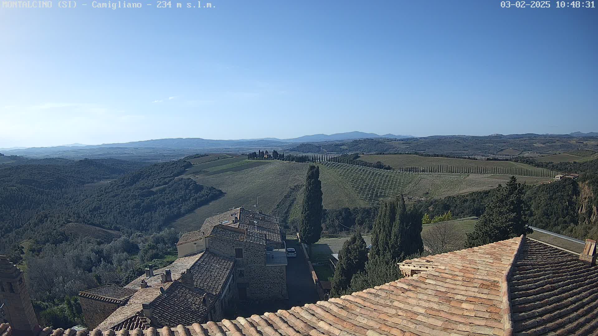 A view from a terracotta roof shows a clear blue sky over rolling hills dotted with cypress trees and stone buildings.