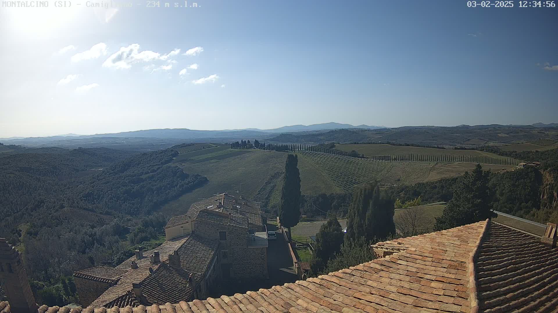 A sunny day reveals a Tuscan landscape of rolling hills, cypress trees, and terracotta rooftops, with distant mountains visible under a mostly clear sky.