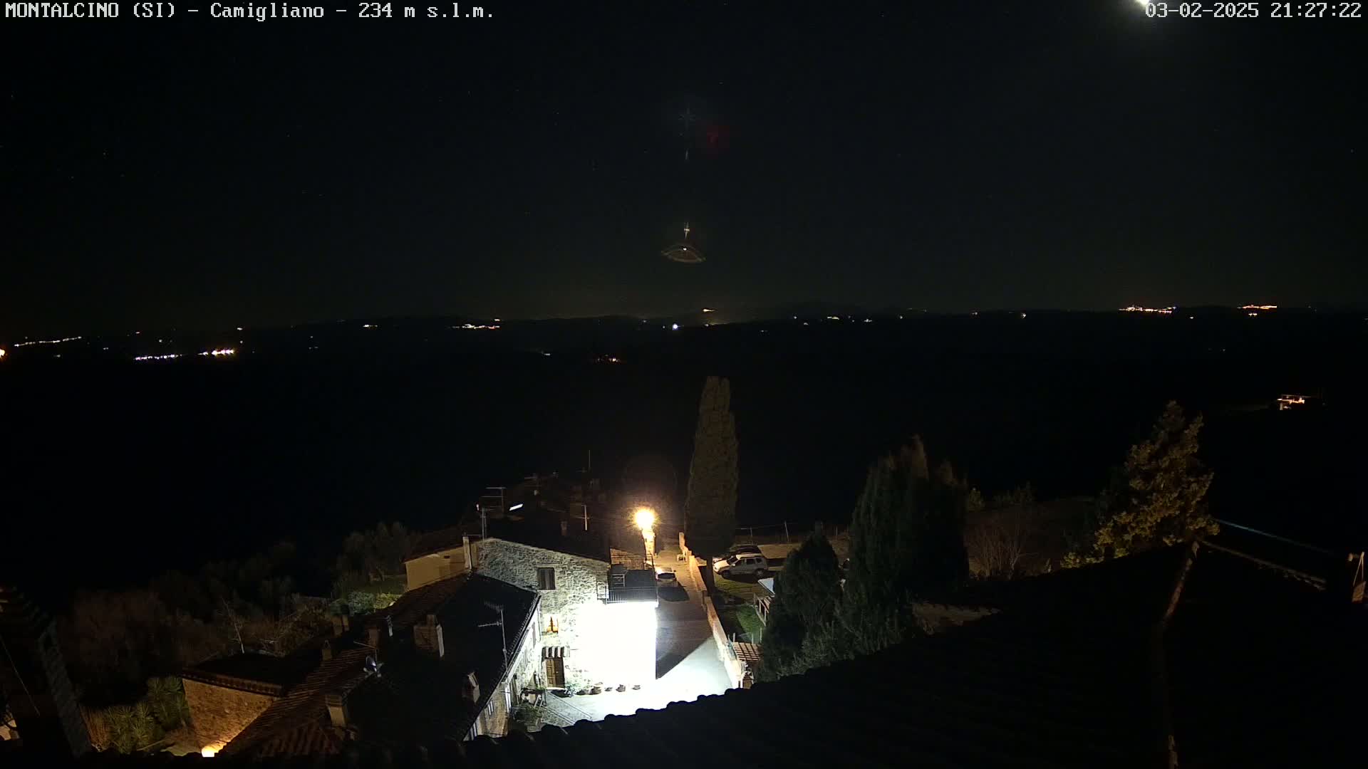 A nighttime, high-angle view shows a village with a brightly lit building and a dark, starlit sky with a faint, possibly meteoric, light trail.