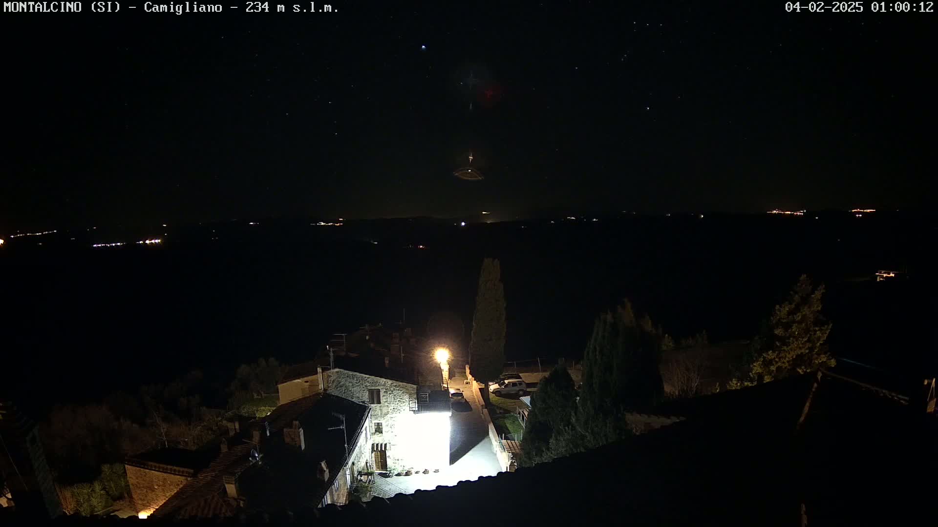 A nighttime view of a small village under a starry sky, with distant lights visible on a hillside.