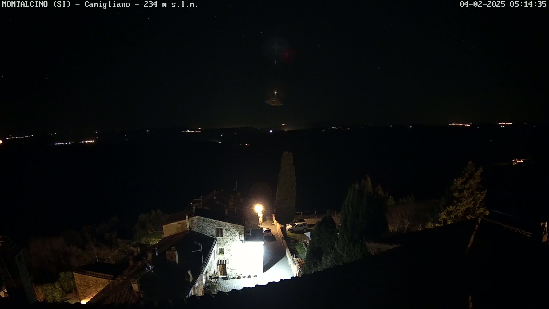 A nighttime, high-angle view of a stone building illuminated by a bright light, situated in a village overlooking a dark landscape speckled with distant lights under a clear, starry sky.