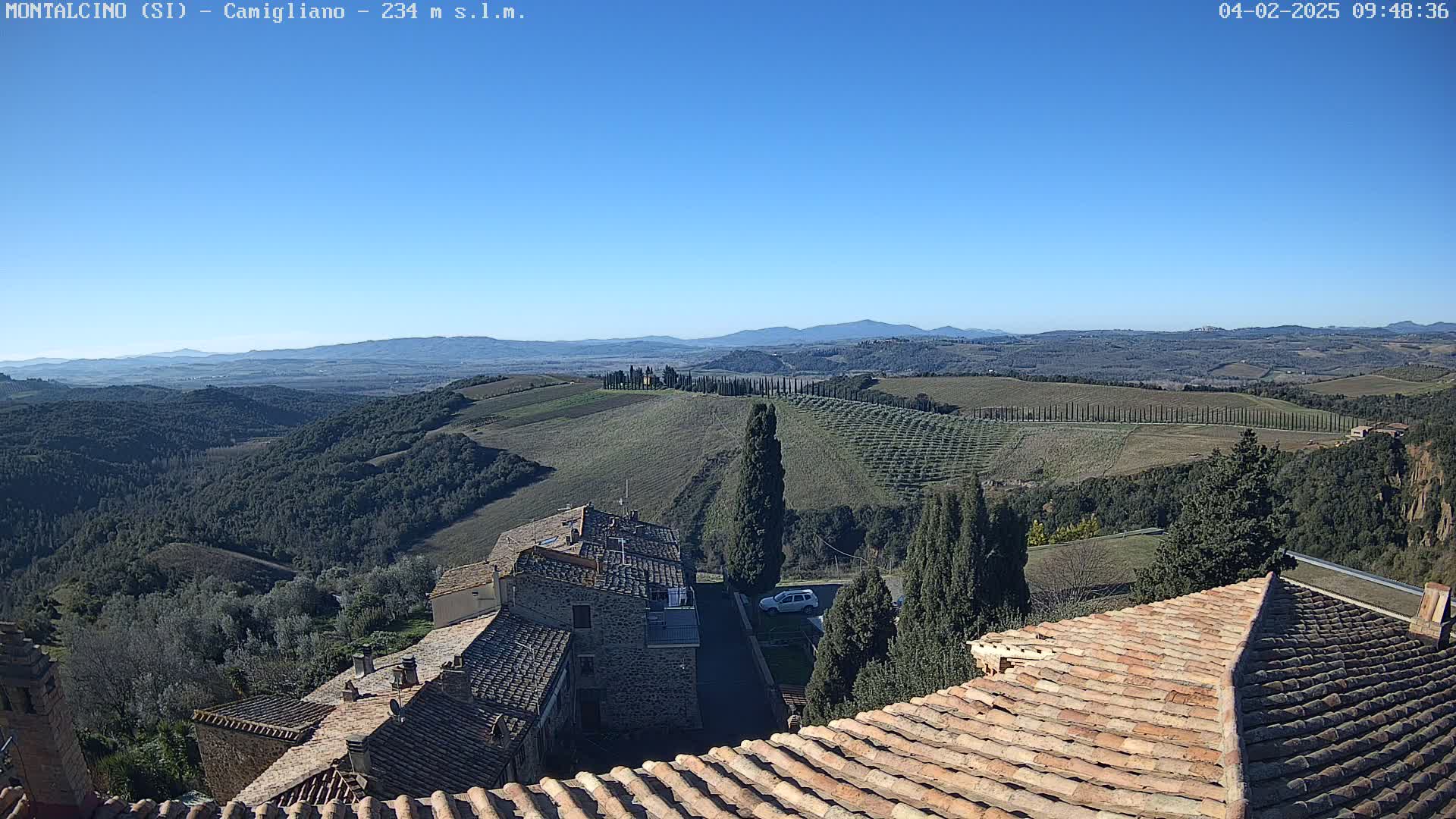 A clear, sunny day reveals a picturesque Tuscan landscape of rolling hills, vineyards, and cypress trees, viewed from atop a village of terracotta-roofed buildings.