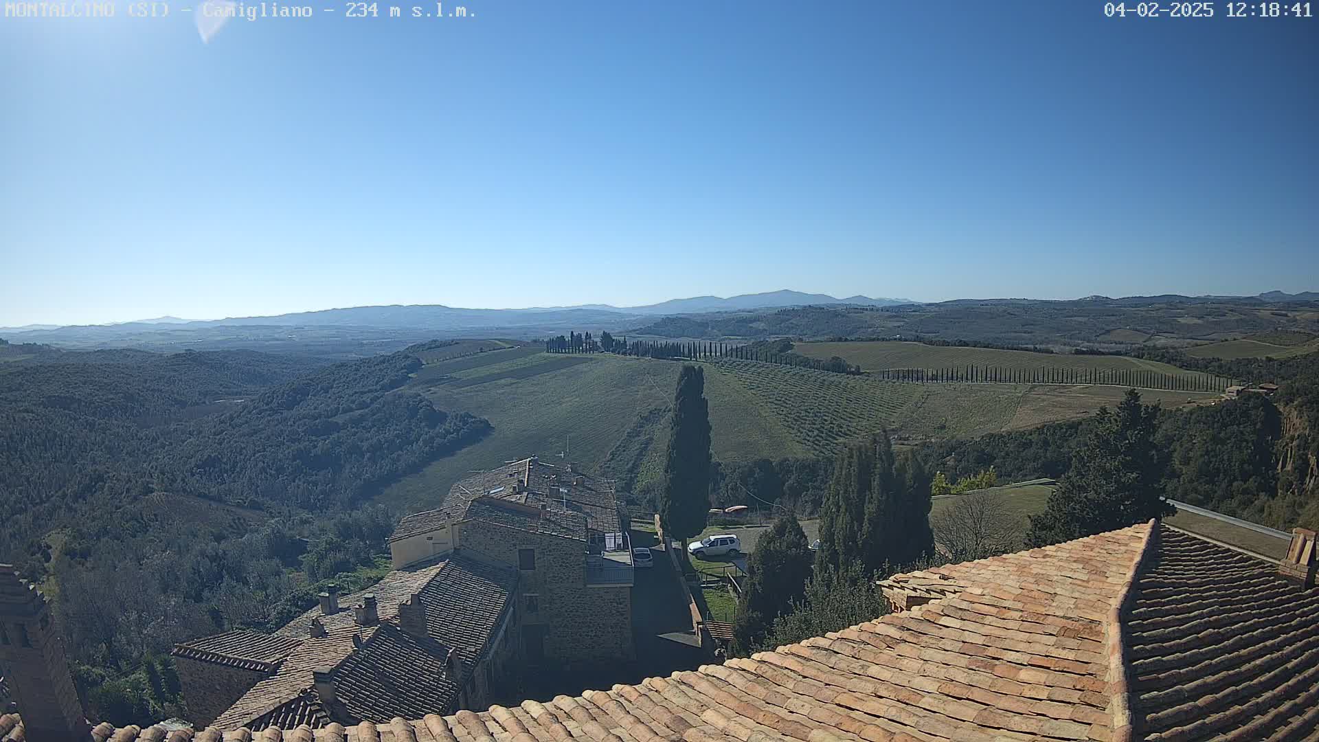 A view from a terracotta roof shows a sunny, clear day overlooking a rolling landscape of green hills, vineyards, and cypress trees, with distant mountains under a bright blue sky.