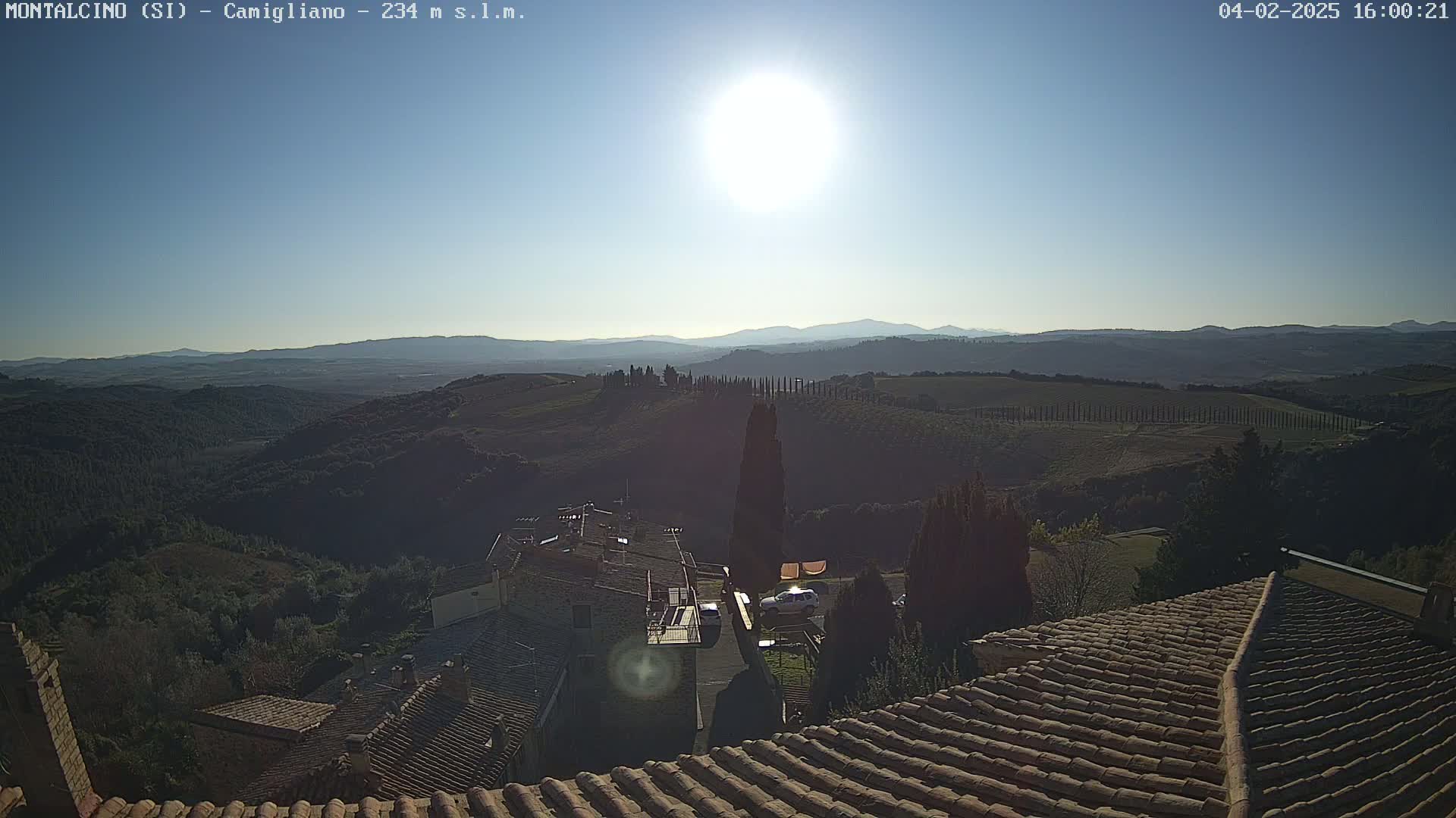 A sunlit view from a rooftop shows rolling hills, vineyards, and a village, under a clear blue sky.