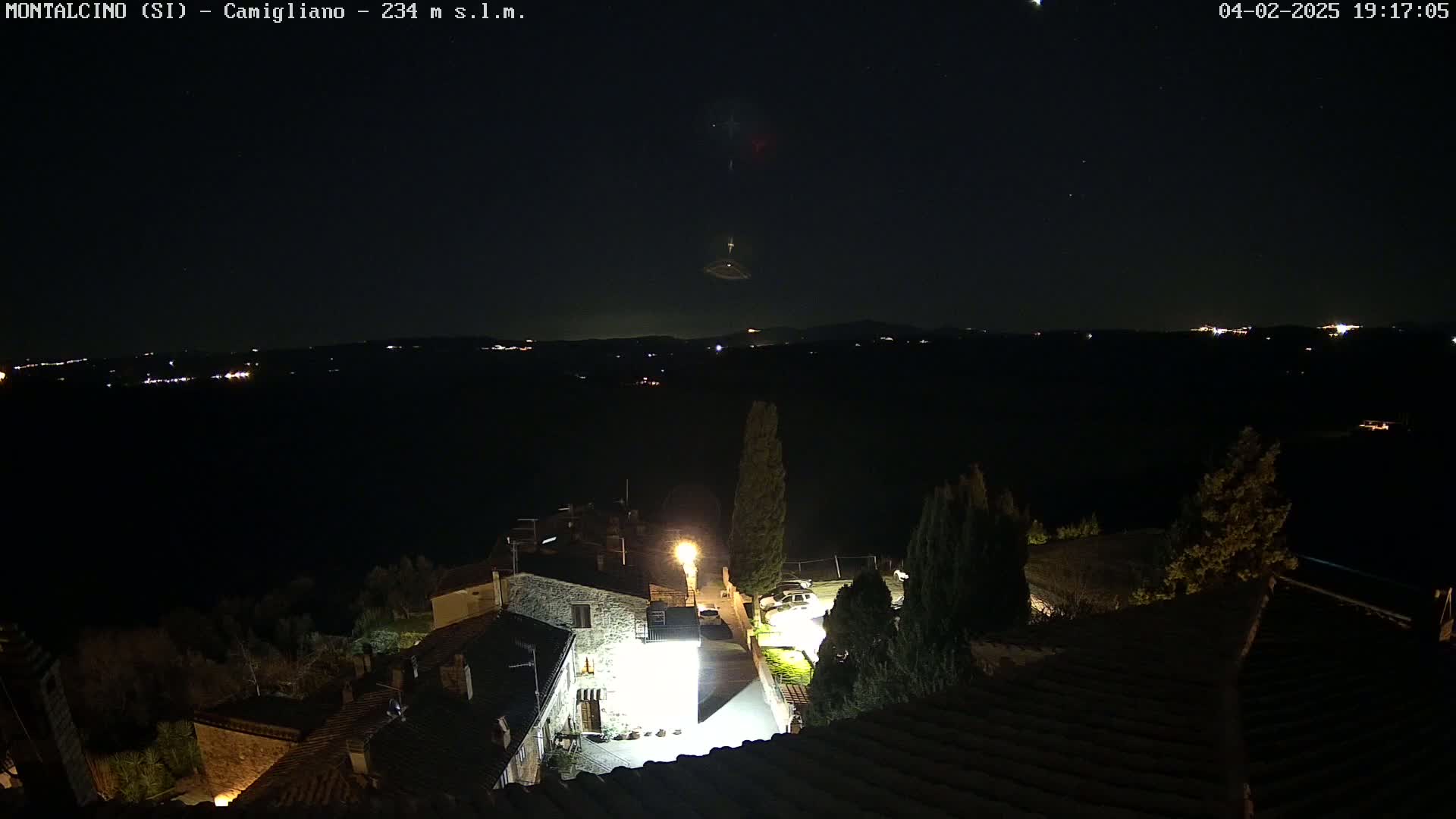 A nighttime view shows a small village with dimly lit stone buildings under a clear, dark sky dotted with stars and a faint light anomaly.