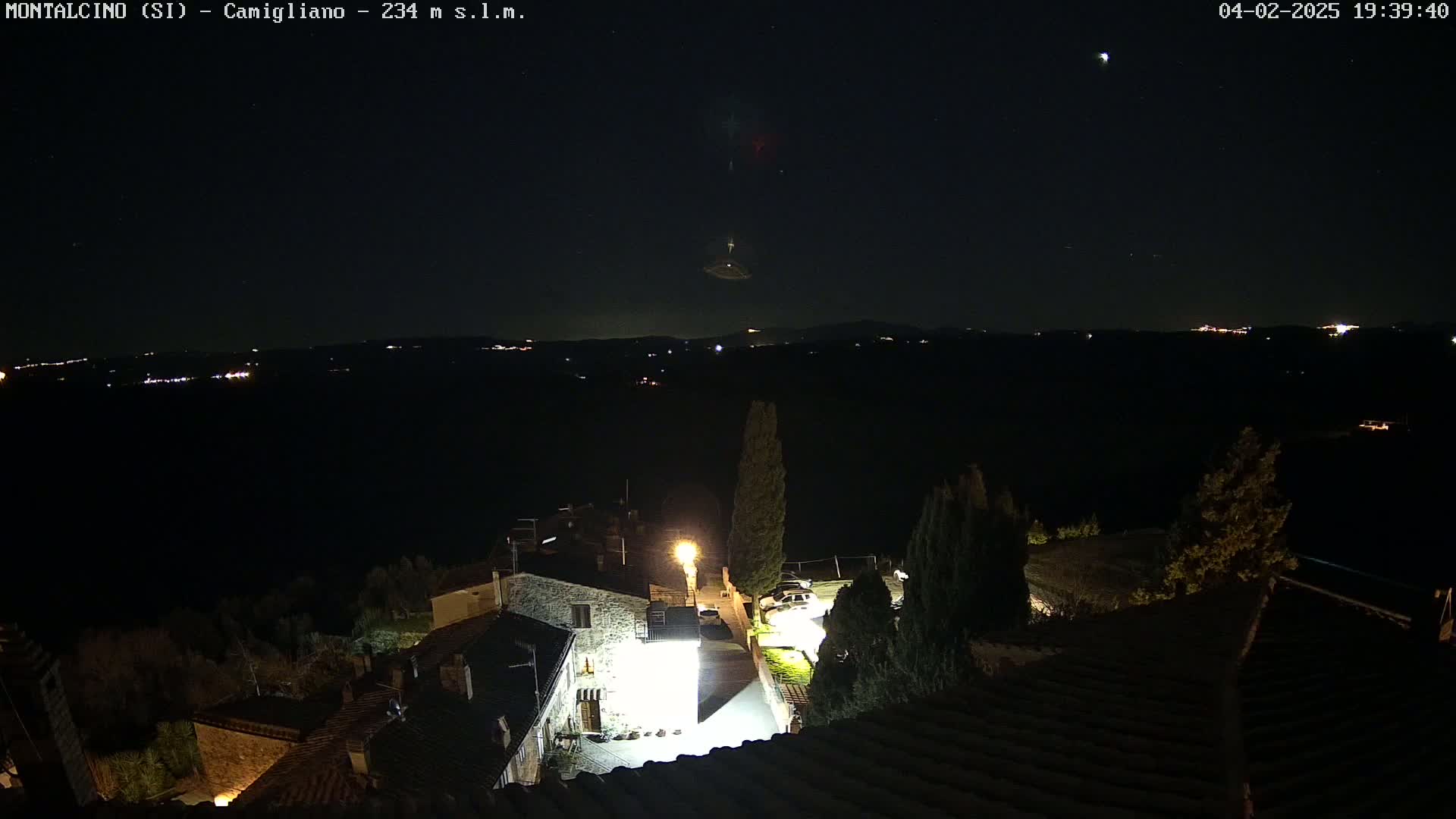 A nighttime view of a village illuminated by artificial lights, under a clear, starry sky with a few distant lights visible on the horizon.
