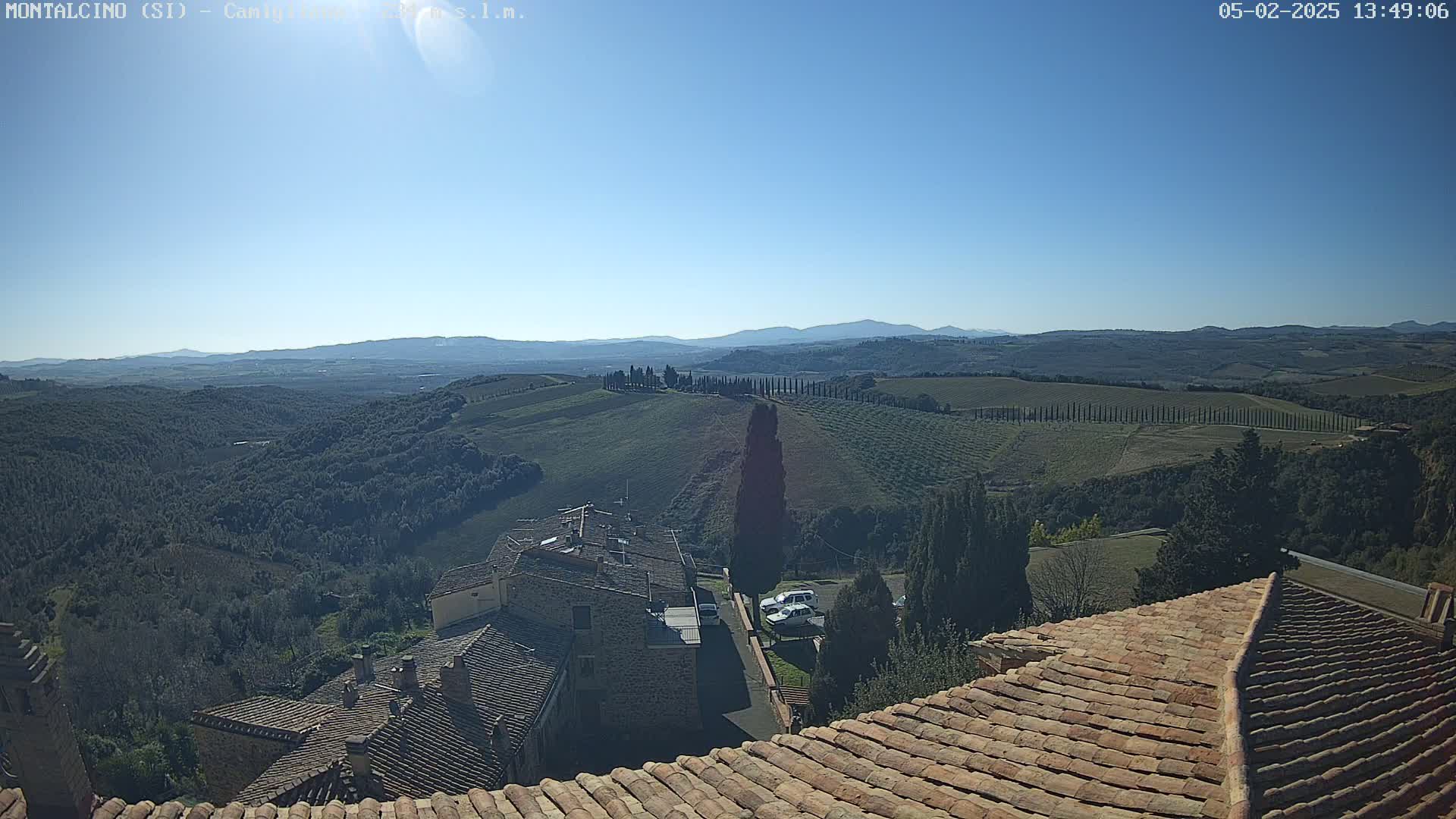 A sunny view of rolling hills and farmland dotted with trees, overlooked by the terracotta roofs of buildings.