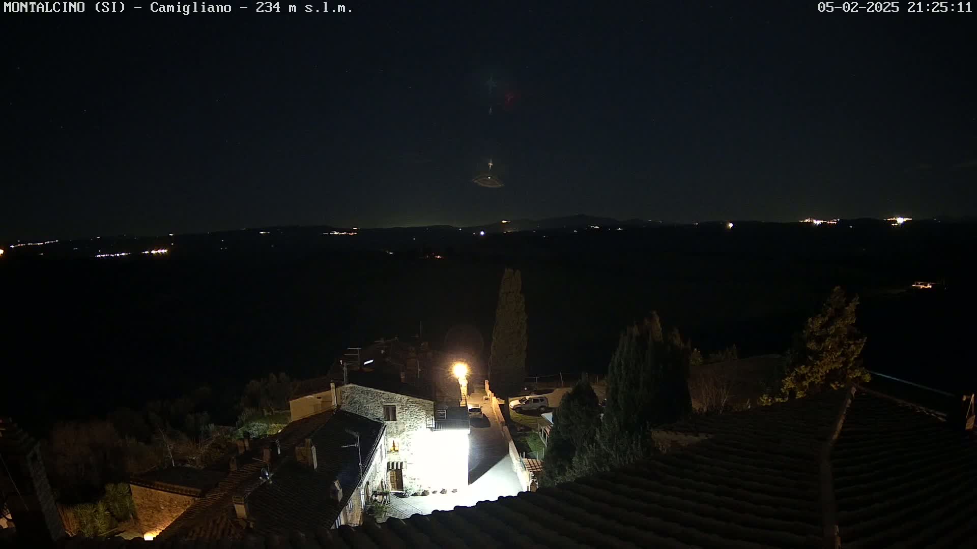 A nighttime, high-angle view of a small village nestled in a dark landscape under a clear, starry sky, with distant lights visible on the horizon.
