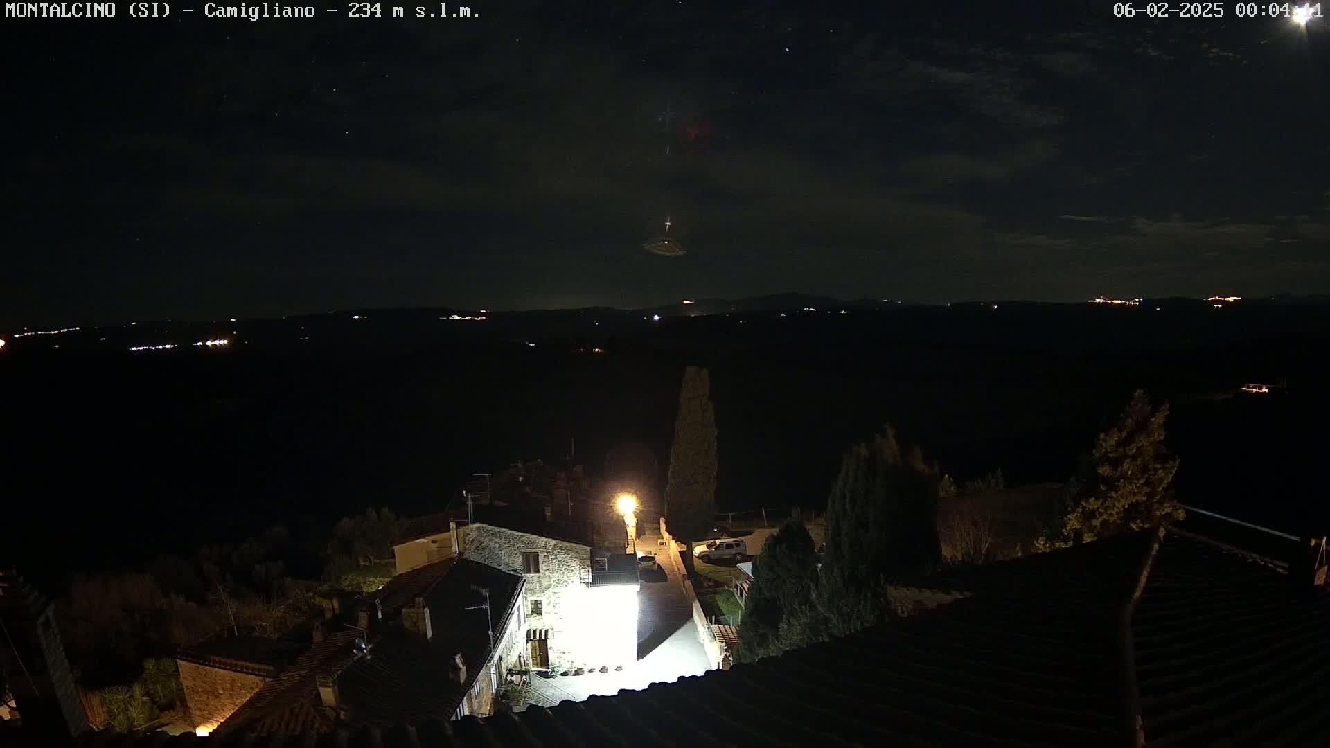 A nighttime view of a small village, illuminated by a few lights, overlooks a dark landscape dotted with distant lights under a mostly clear, starry sky.