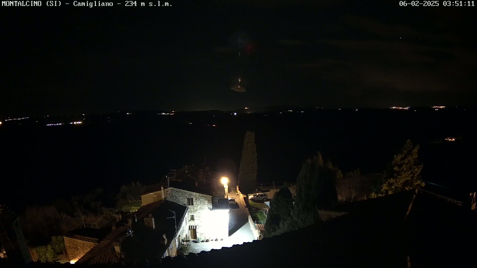 A nighttime, high-angle view shows a small village illuminated by streetlights, with a distant, faintly lit landscape under a mostly clear, dark sky.