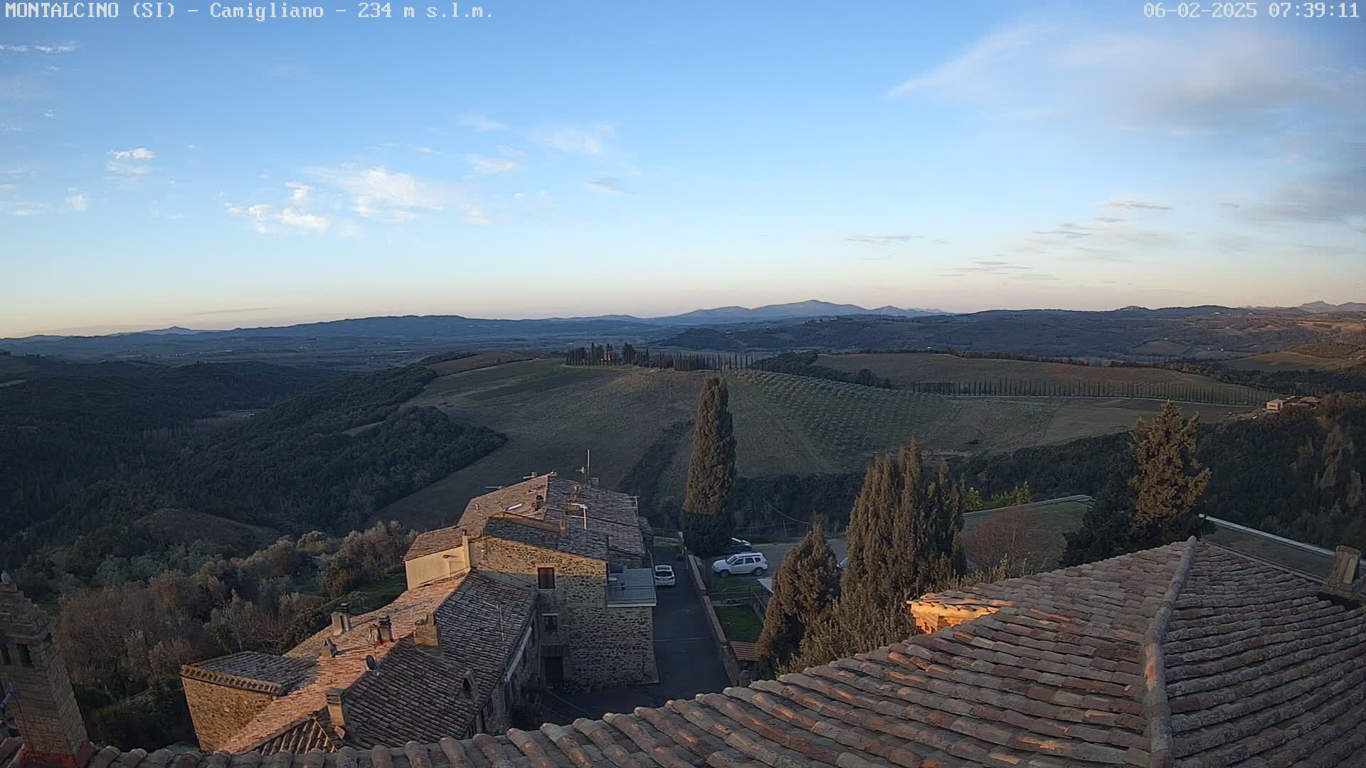 Stone rooftops overlook a sunlit valley of rolling hills and farmland under a mostly clear, pale blue sky.