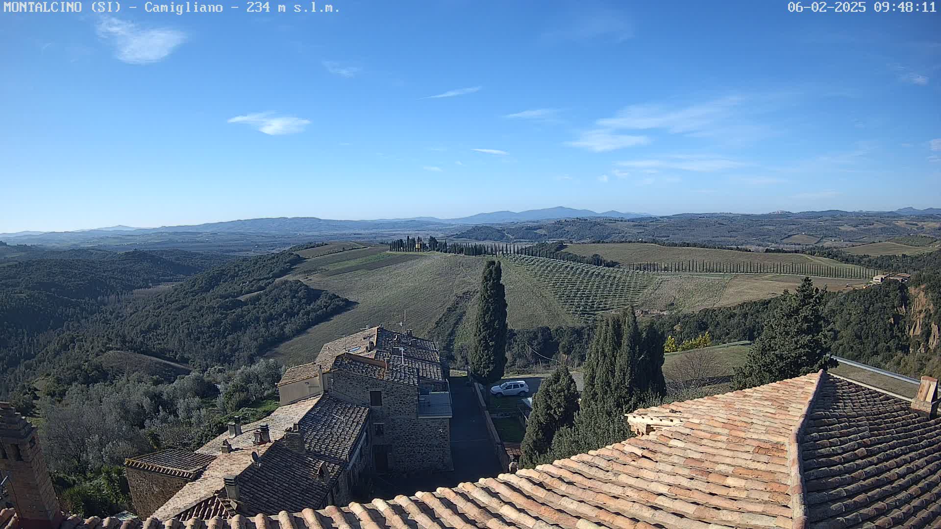A clear blue sky overlooks a Tuscan landscape of rolling hills, vineyards, and a village with terracotta rooftops.