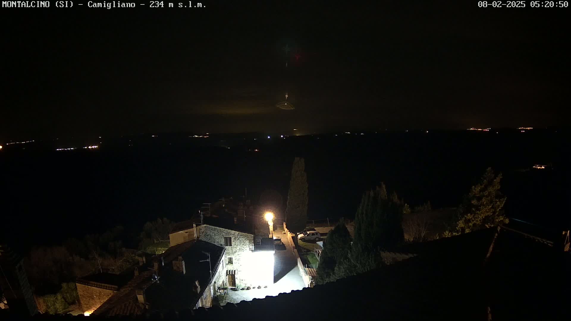 A nighttime view of a village, illuminated by streetlights, overlooks a dark, distant landscape with faintly visible lights under a clear night sky.