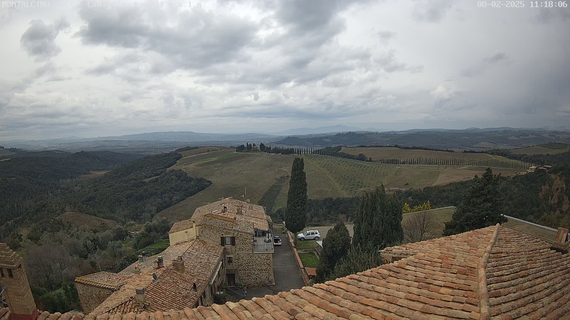 Overcast skies reveal a sprawling Tuscan landscape of rolling hills, cypress trees, and terracotta-roofed buildings.