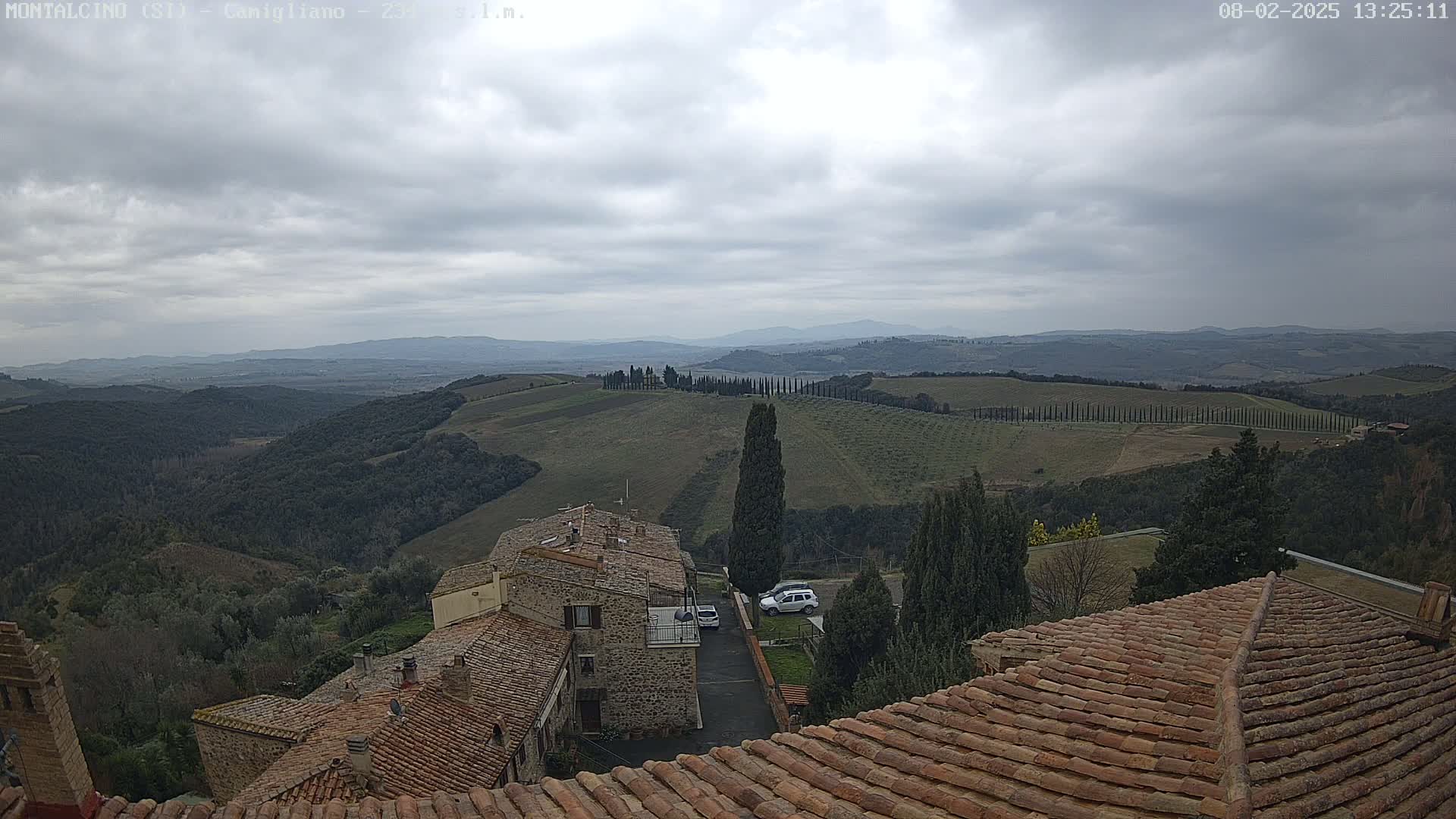 Overcast skies reveal a view of rolling hills dotted with cypress trees and stone buildings with terracotta roofs.