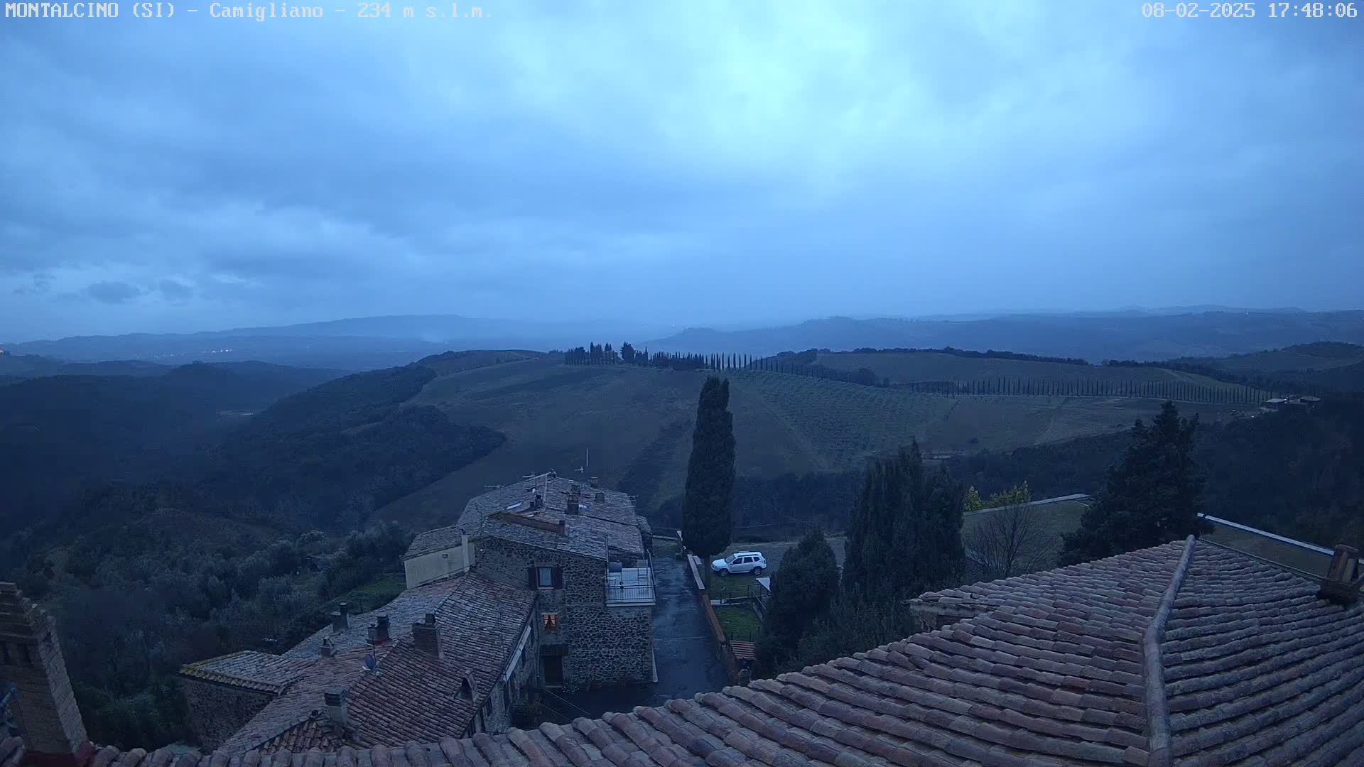 A view from a rooftop shows a twilight scene over rolling hills and a small village, under a dusky blue sky.