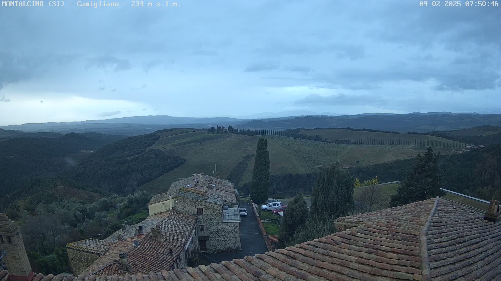 Overcast skies and rolling hills of a rural landscape are viewed from atop terracotta rooftops of stone buildings.