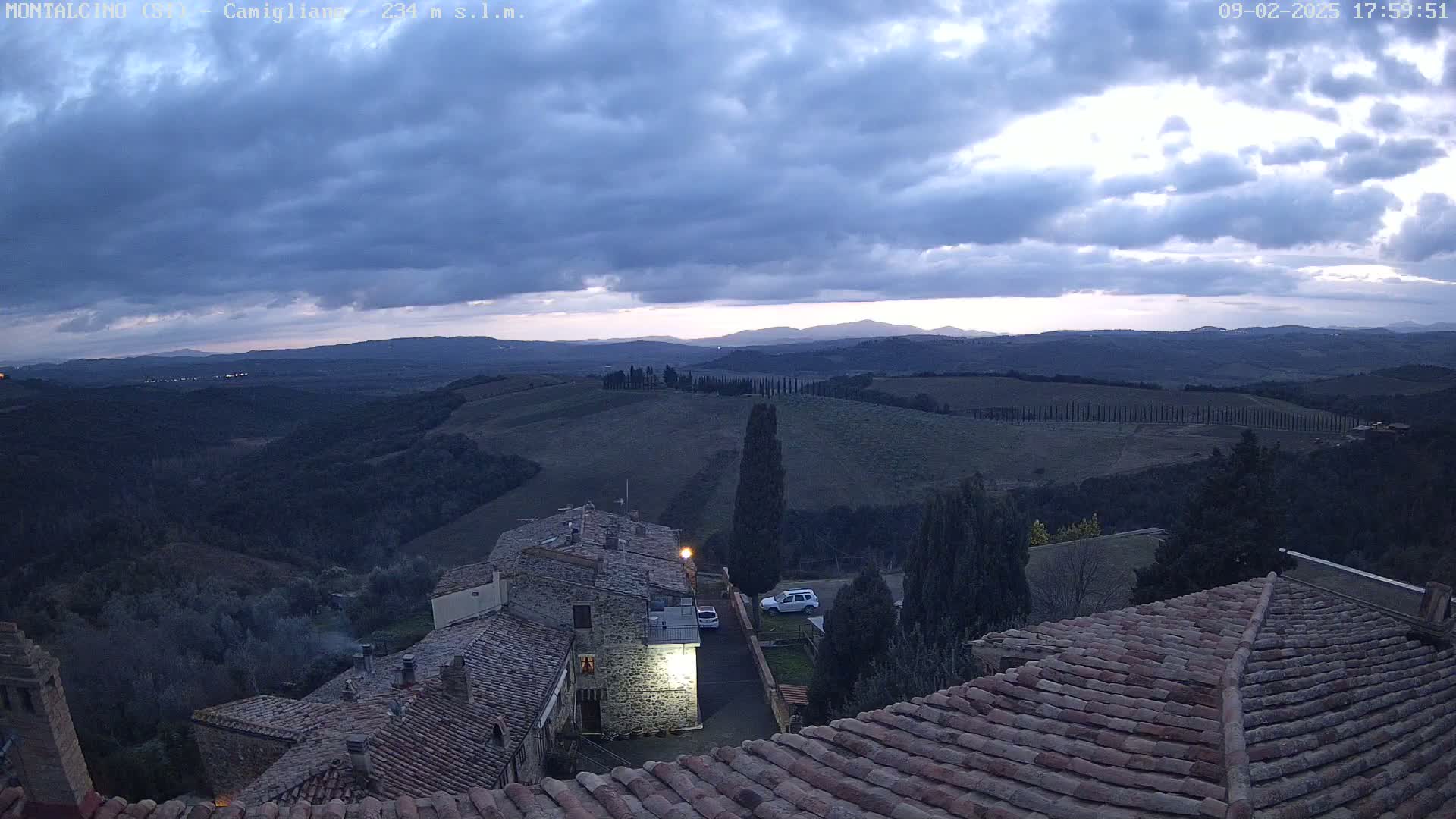 A view from a rooftop shows a hilly landscape under a cloudy twilight sky, with stone buildings and a few cars visible in the foreground.