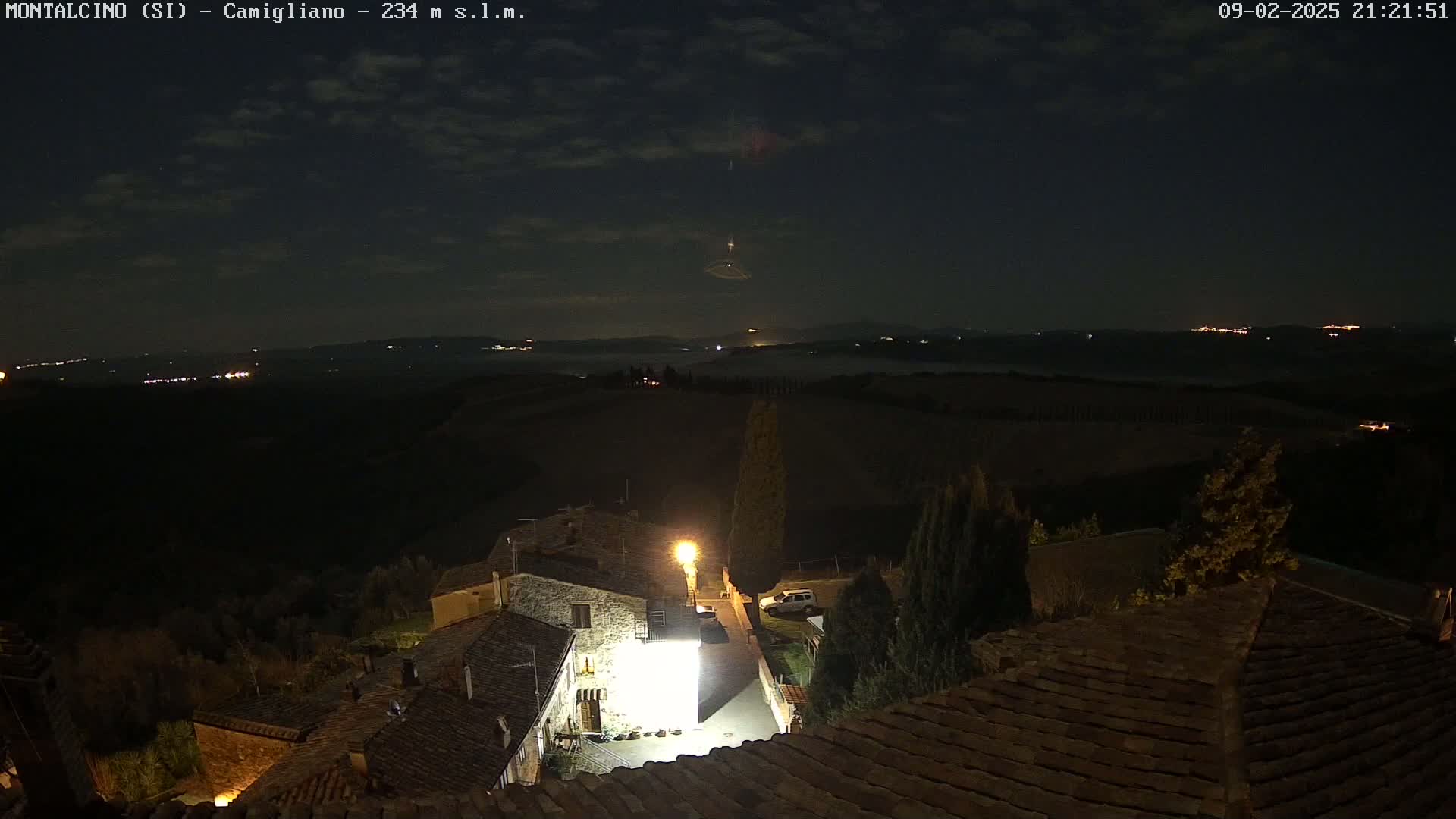 A nighttime view of a village with low-lying fog in the distance under a partly cloudy sky.