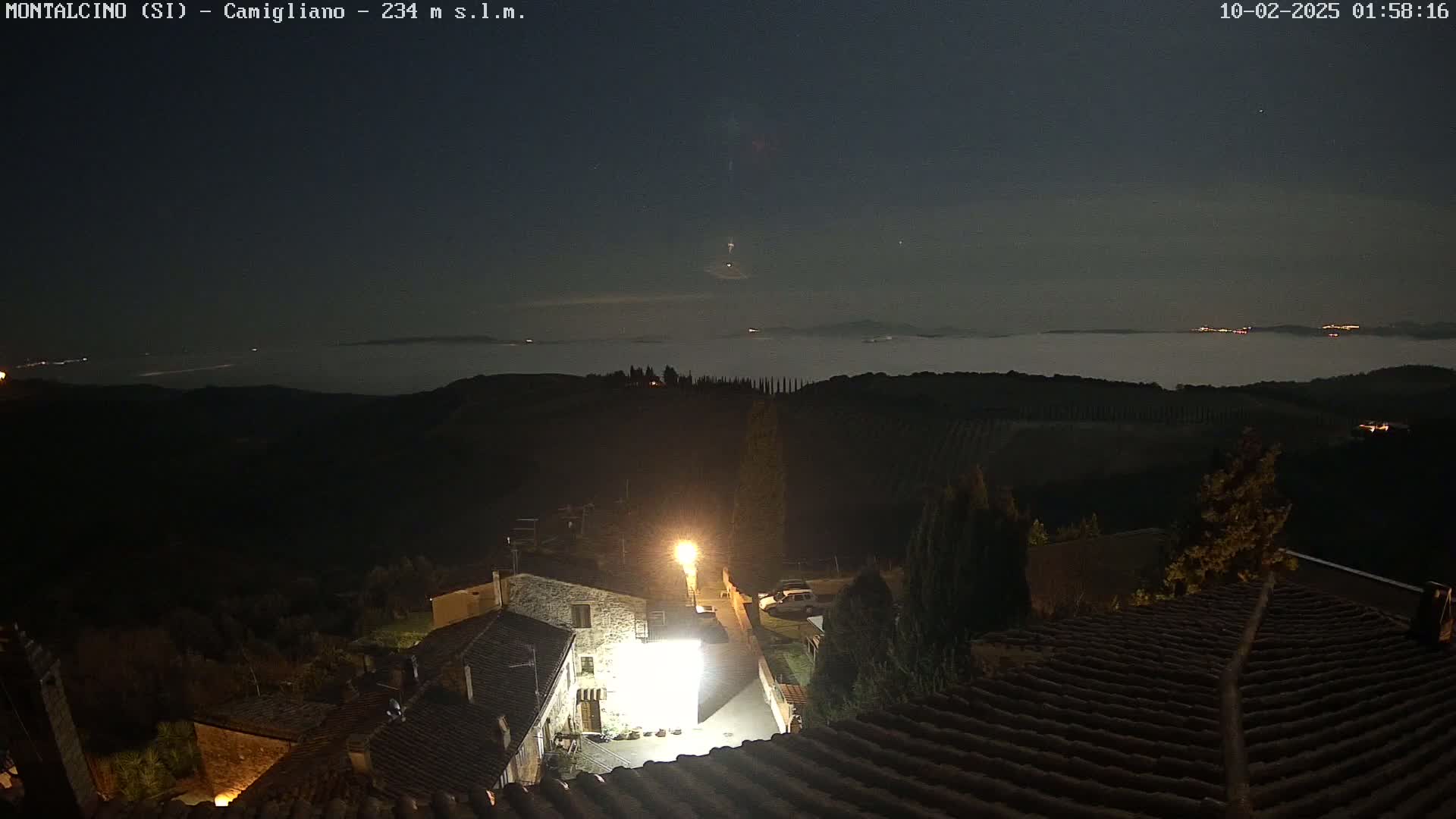 A nighttime view from a hilltop shows a low-lying fog over a valley, with a small village illuminated by a single streetlight in the foreground under a clear, dark sky.