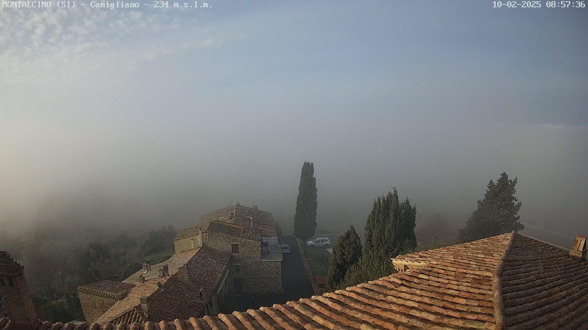 A foggy, hazy view from a rooftop shows several stone buildings with terracotta roofs, a few evergreen trees, and a car parked in a yard.