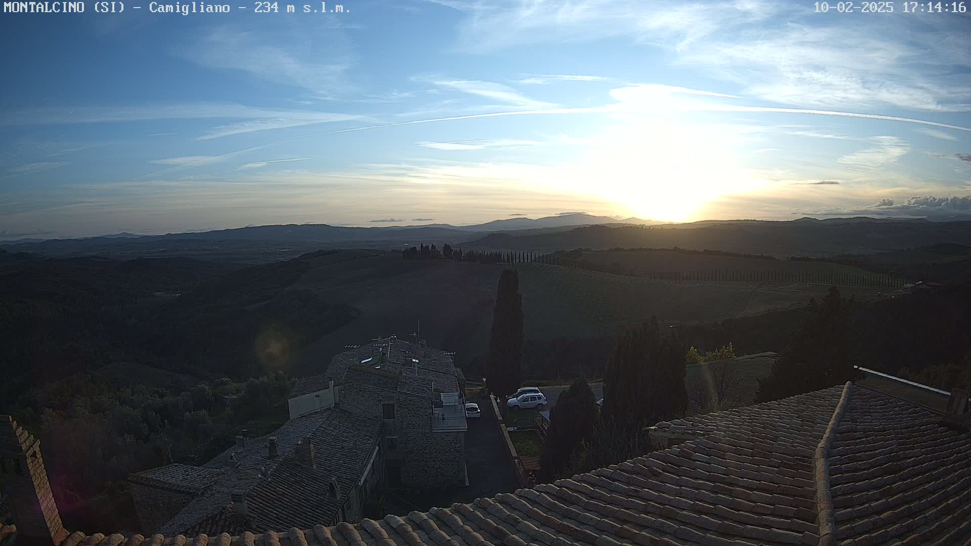 A sunset over rolling hills and a village with terracotta roofs is visible from a high vantage point on a clear day.
