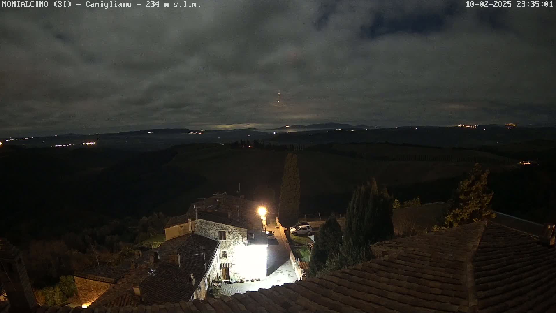 A nighttime view from a rooftop shows a stone village under a cloudy sky, with distant lights illuminating a rolling landscape.