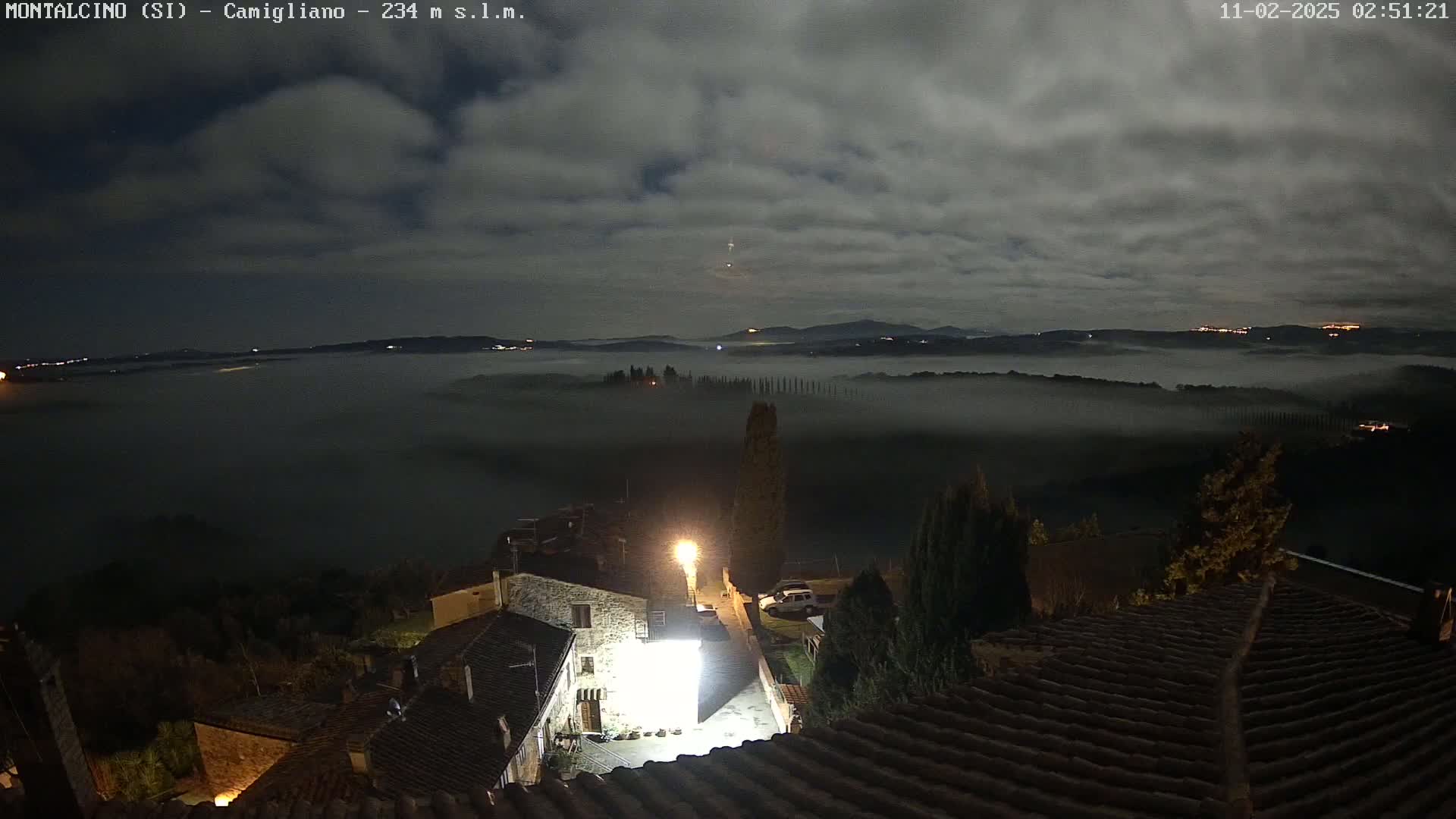 A nighttime view from a hilltop shows a village with low-lying fog obscuring the valley below under a cloudy sky.