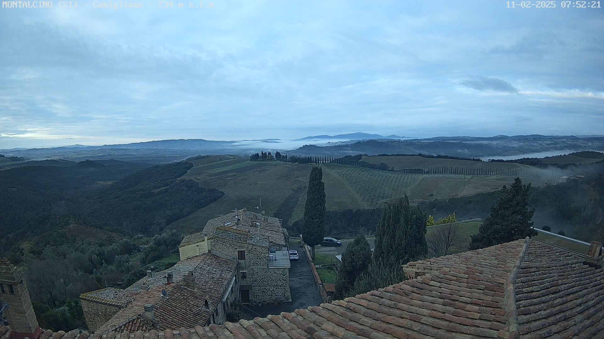 A view from a rooftop shows stone buildings and rolling hills covered in low fog under a cloudy sky.