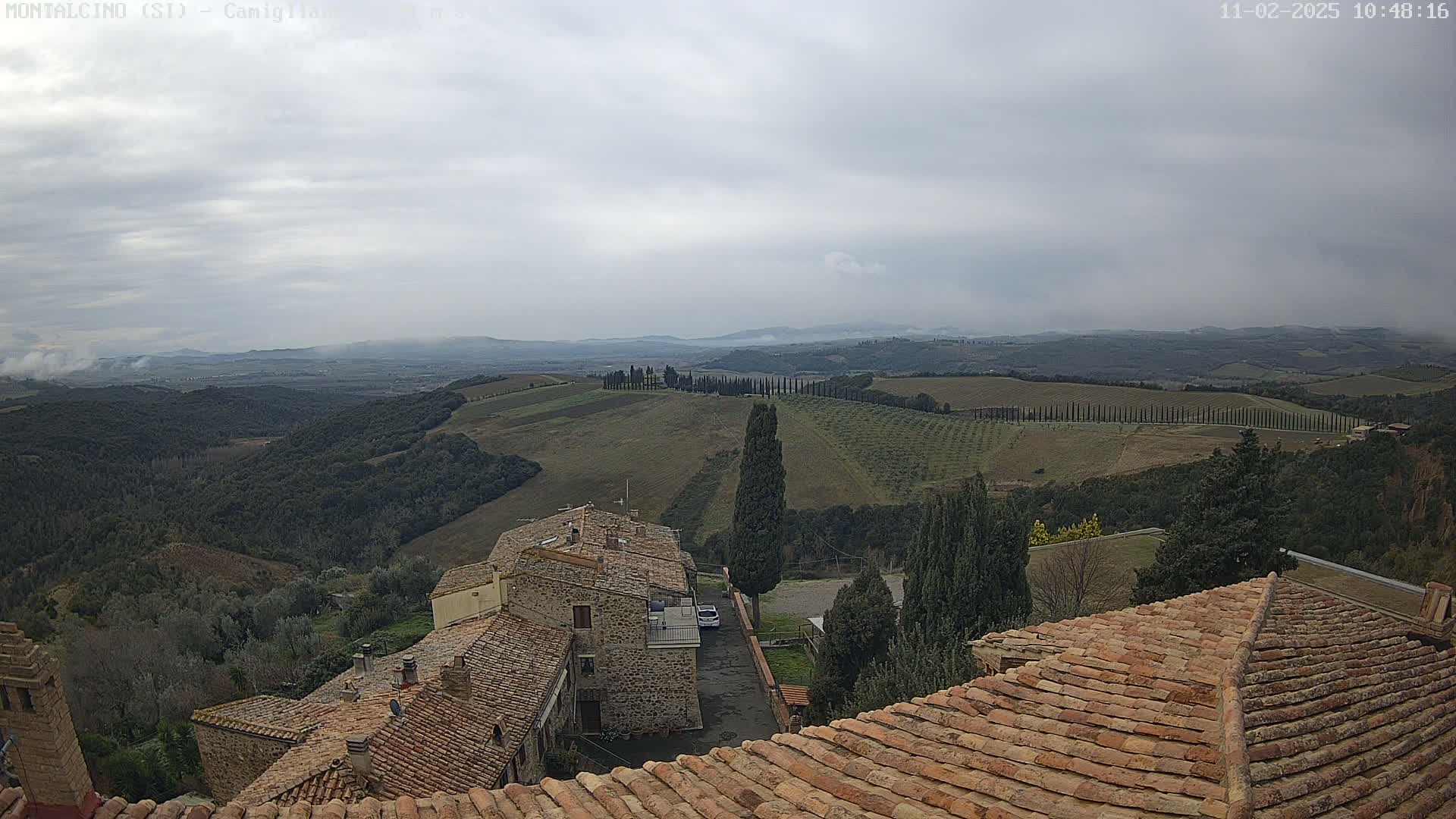 A view from a rooftop shows rolling hills and farmland under an overcast sky, with stone buildings and terracotta roofs in the foreground.