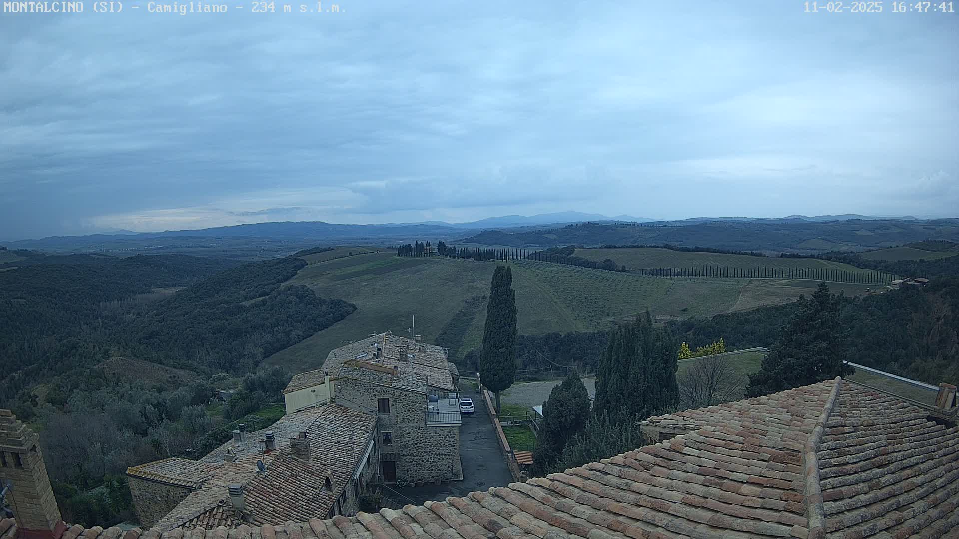 A view from a rooftop shows a cloudy sky over rolling hills and a town with stone buildings and terracotta roofs.