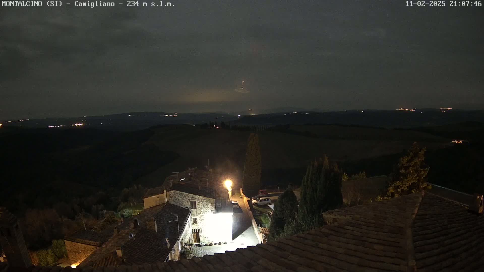 A nighttime view of a village nestled in a hilly landscape under a dark, cloudy sky, with distant lights visible on the horizon.