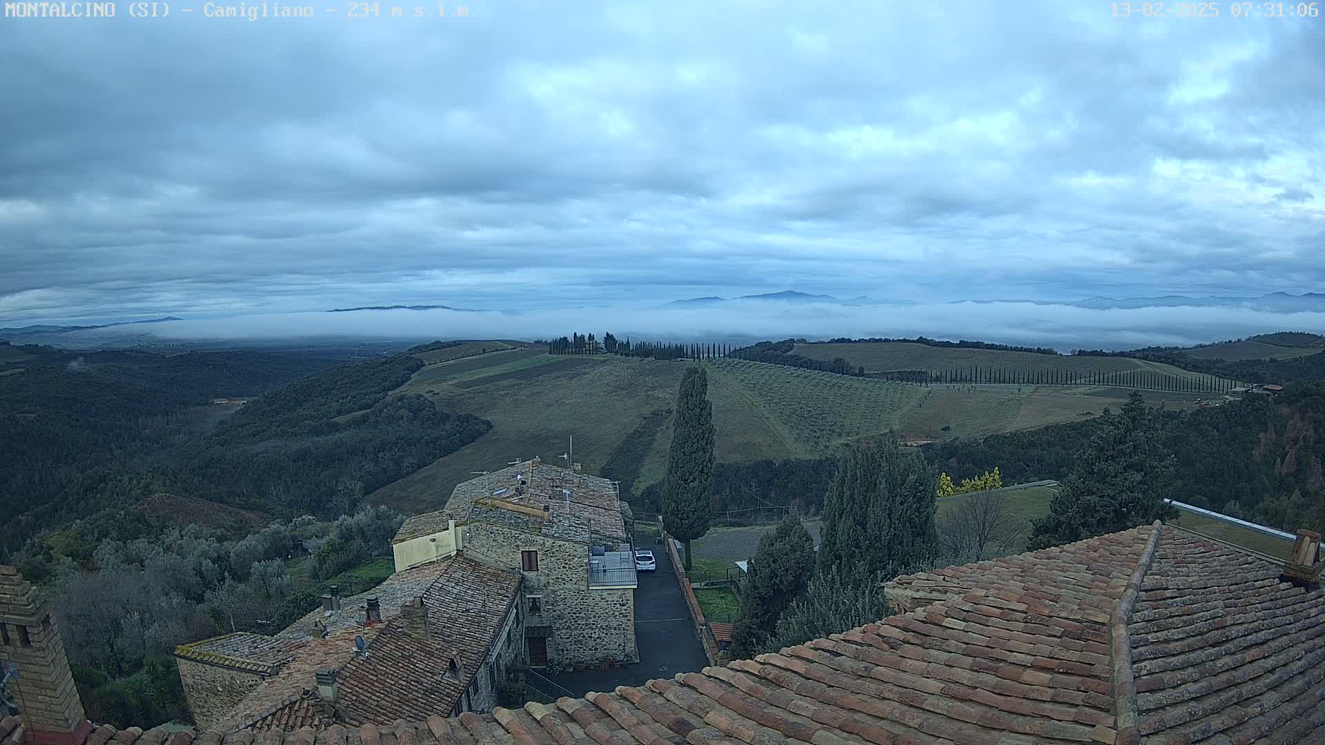 A view from a rooftop shows stone buildings and rolling hills under a cloudy sky with low-lying fog in the distance.