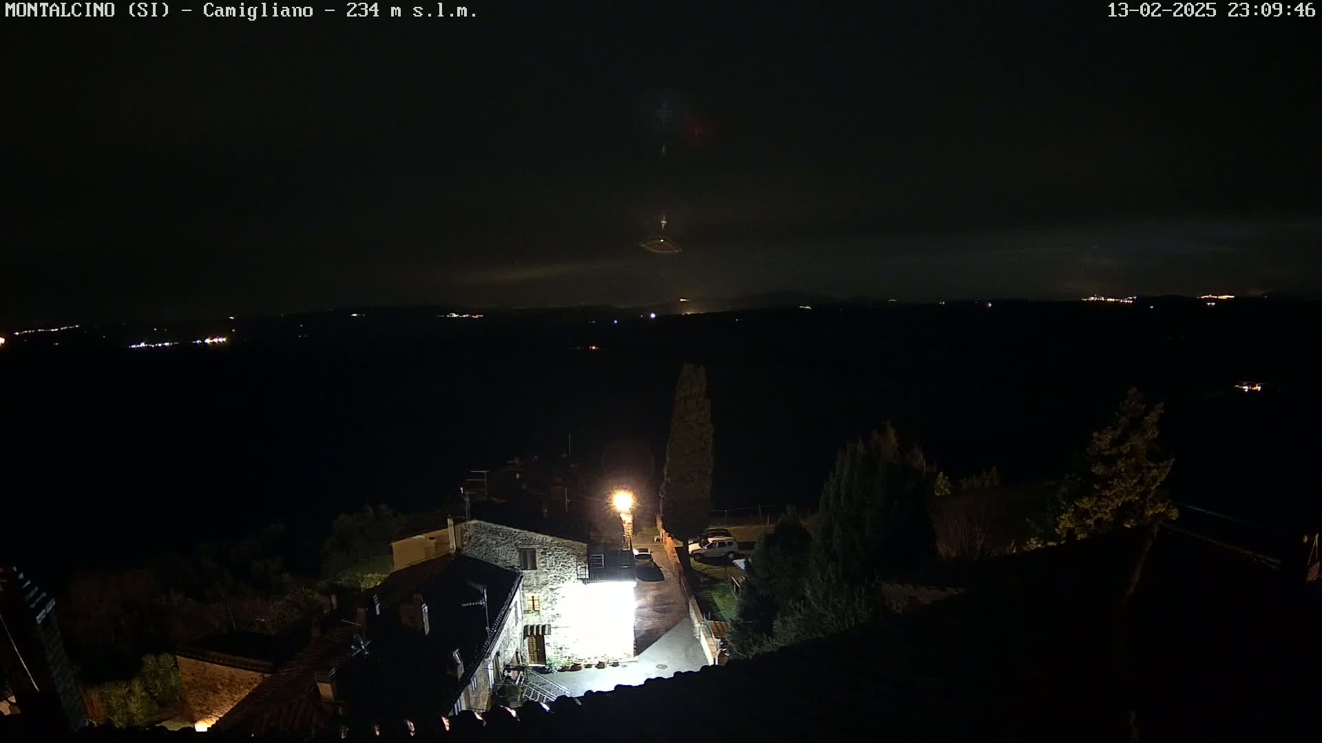 A nighttime view of a small village illuminated by a single bright light, overlooking a dark valley with distant lights under a mostly clear sky.